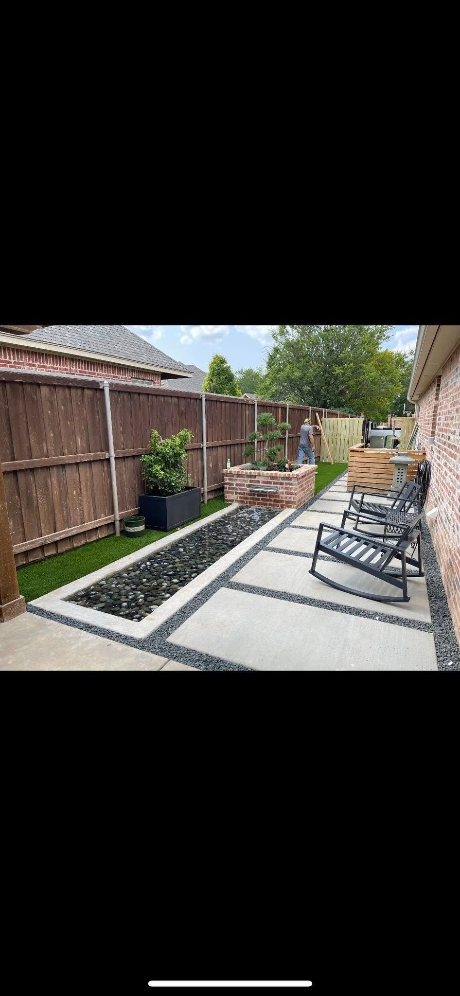 Backyard patio with concrete path, raised flower bed, wooden fence, and outdoor chairs under blue sky