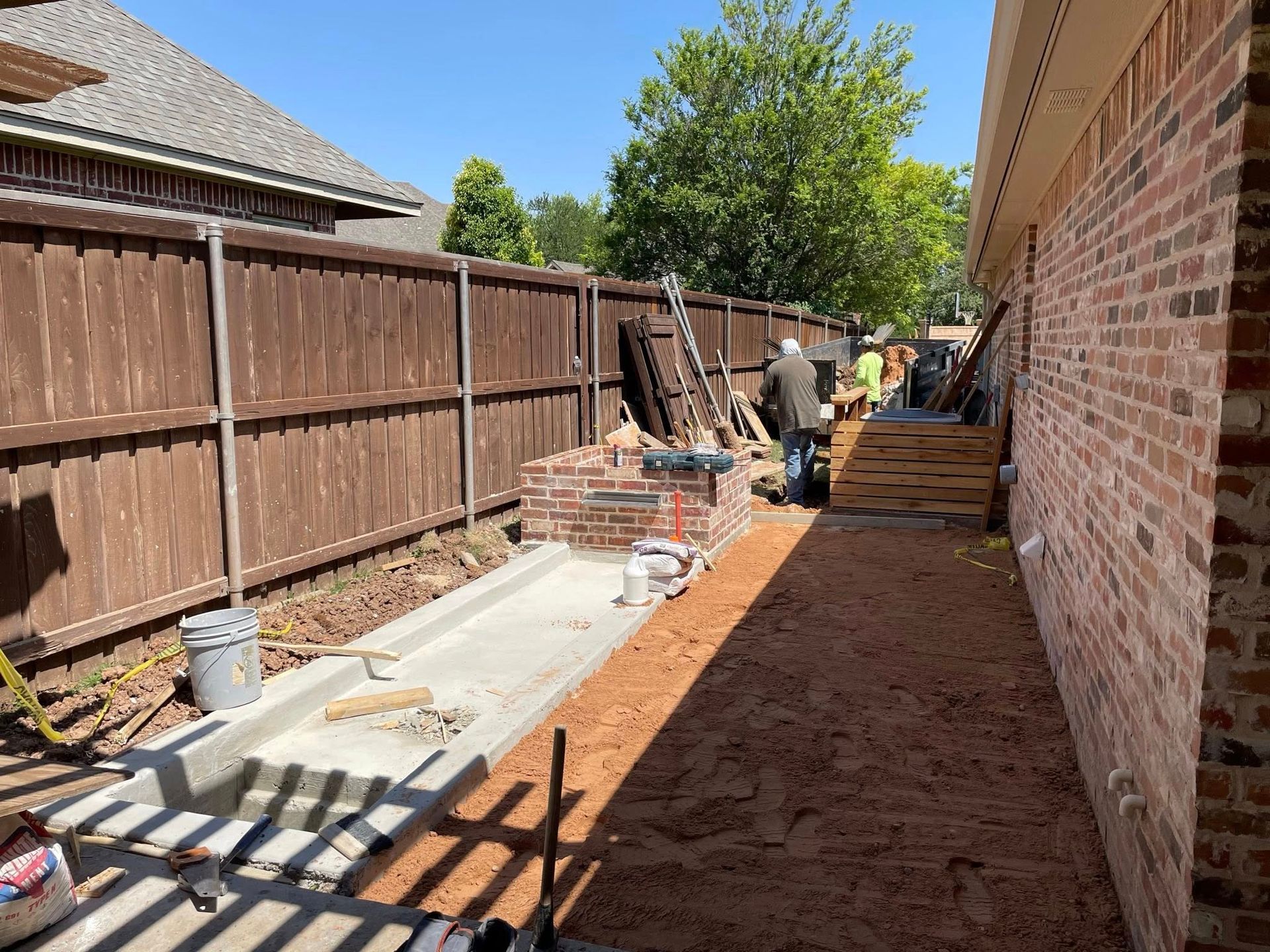 Backyard construction site with brick house, wooden fence, workers, and a concrete walkway under construction