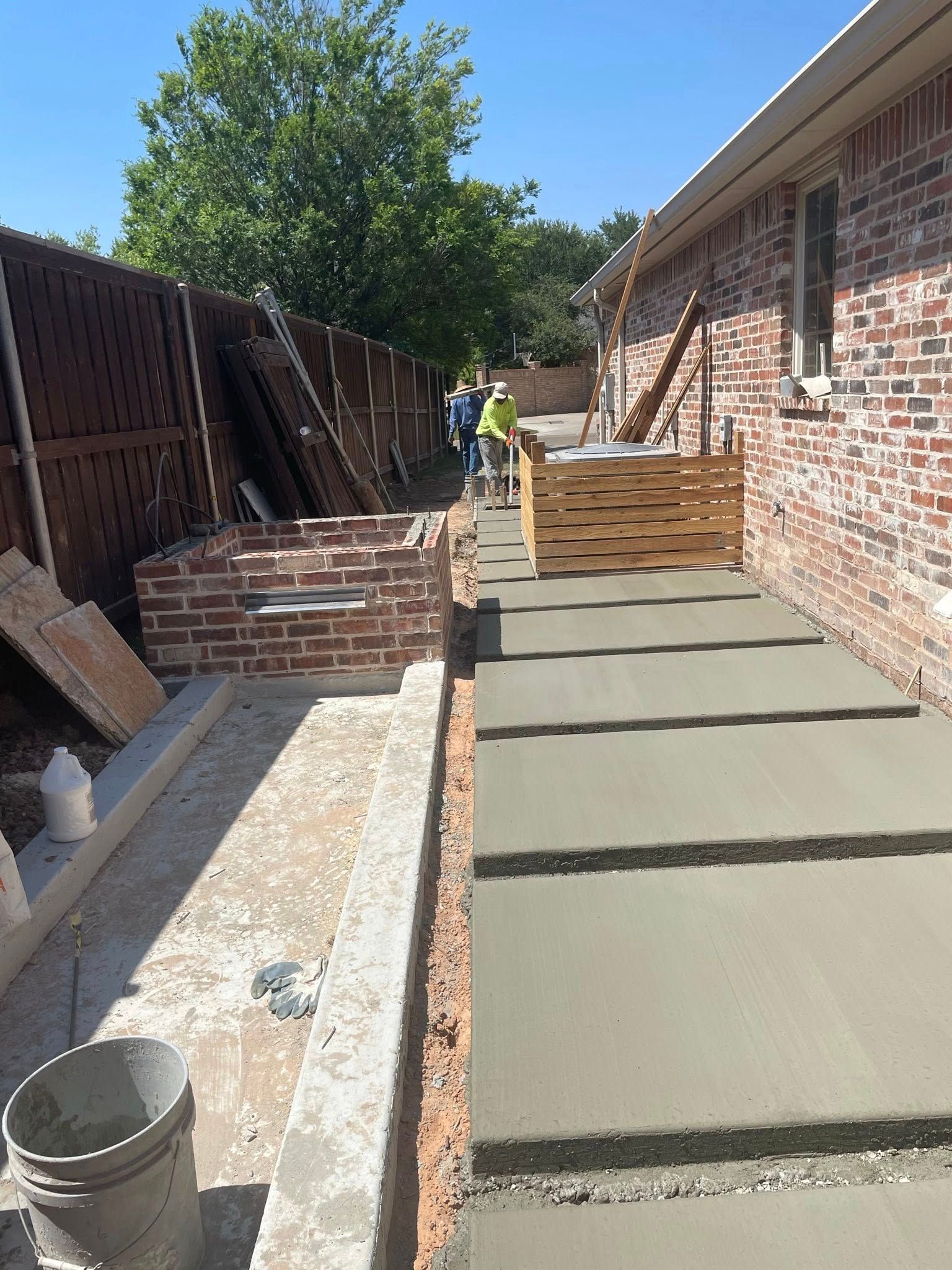 Concrete walkway under construction beside a brick house, with workers and tools in a narrow side yard