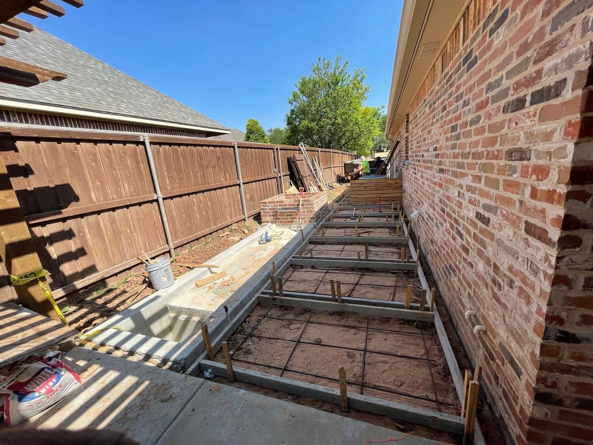 Narrow construction trench beside a brick wall, with steel framing and wooden fence panels along the side.