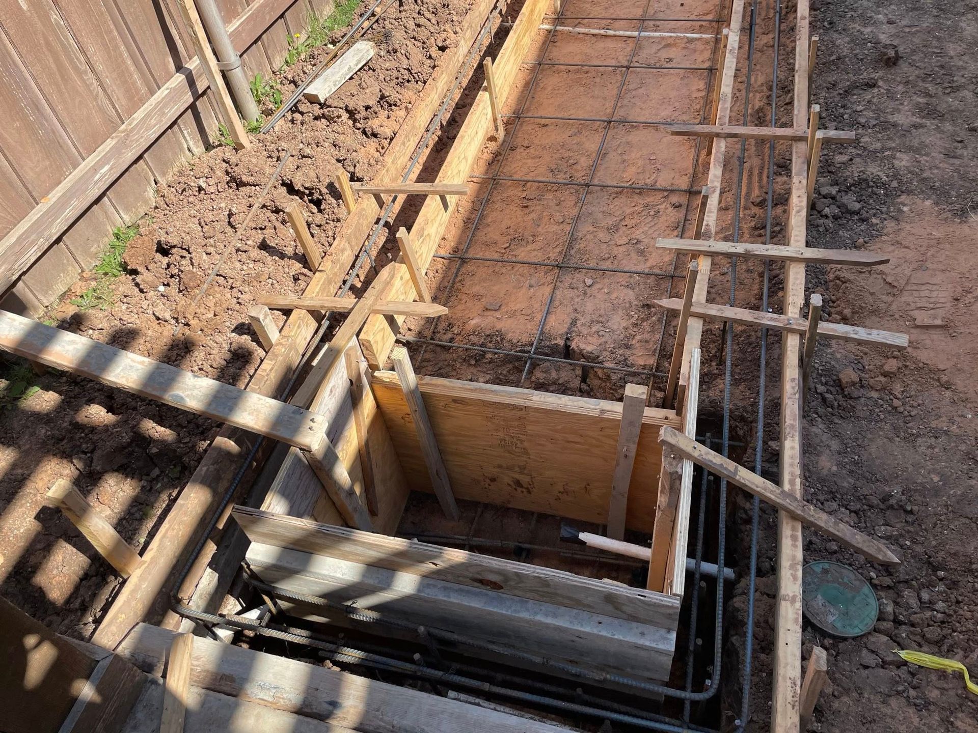 Construction pit with wooden formwork and rebar in a narrow trench beside a fence