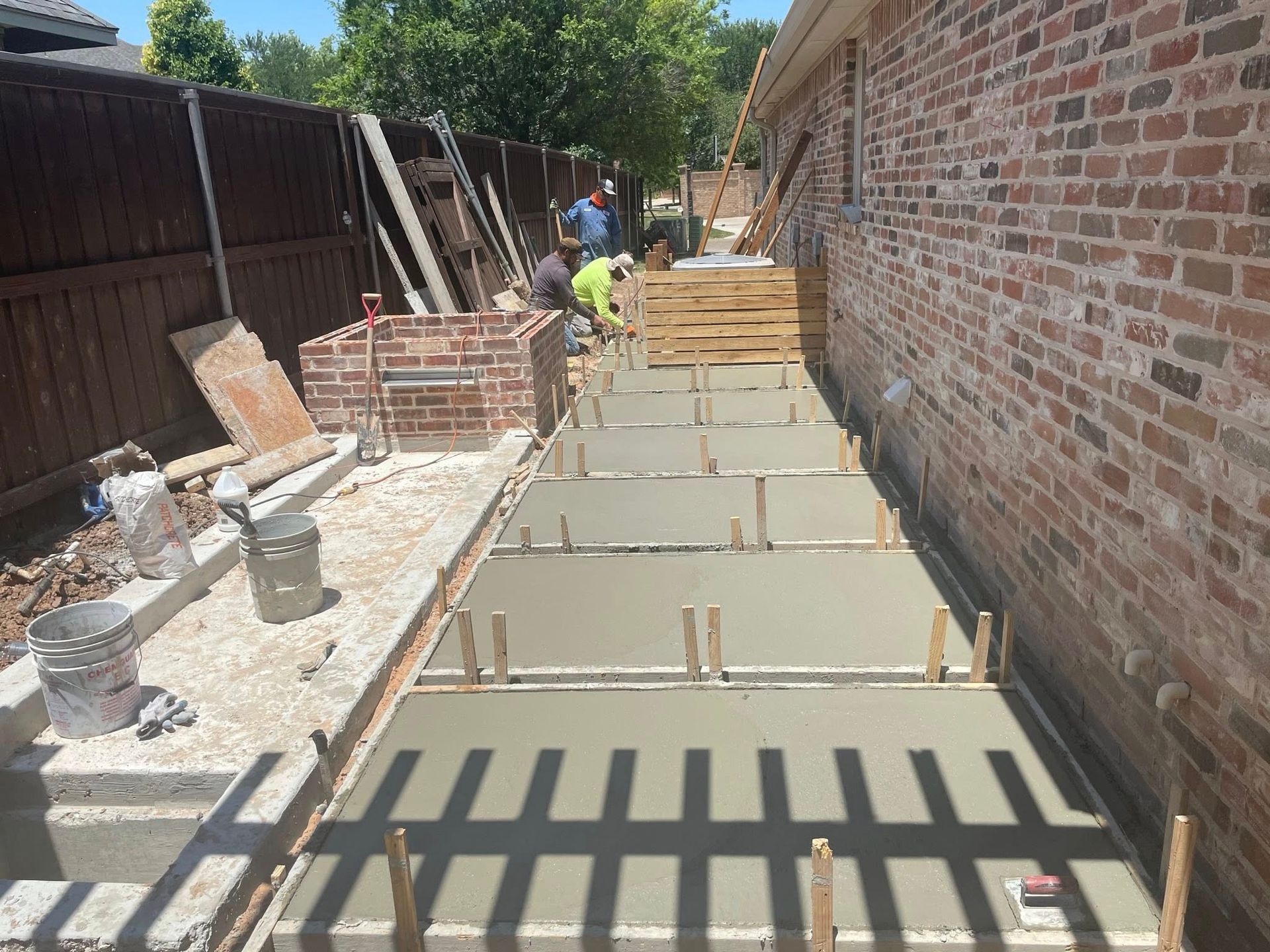 Concrete slab being poured in a narrow backyard beside a brick wall, with workers in the distance.