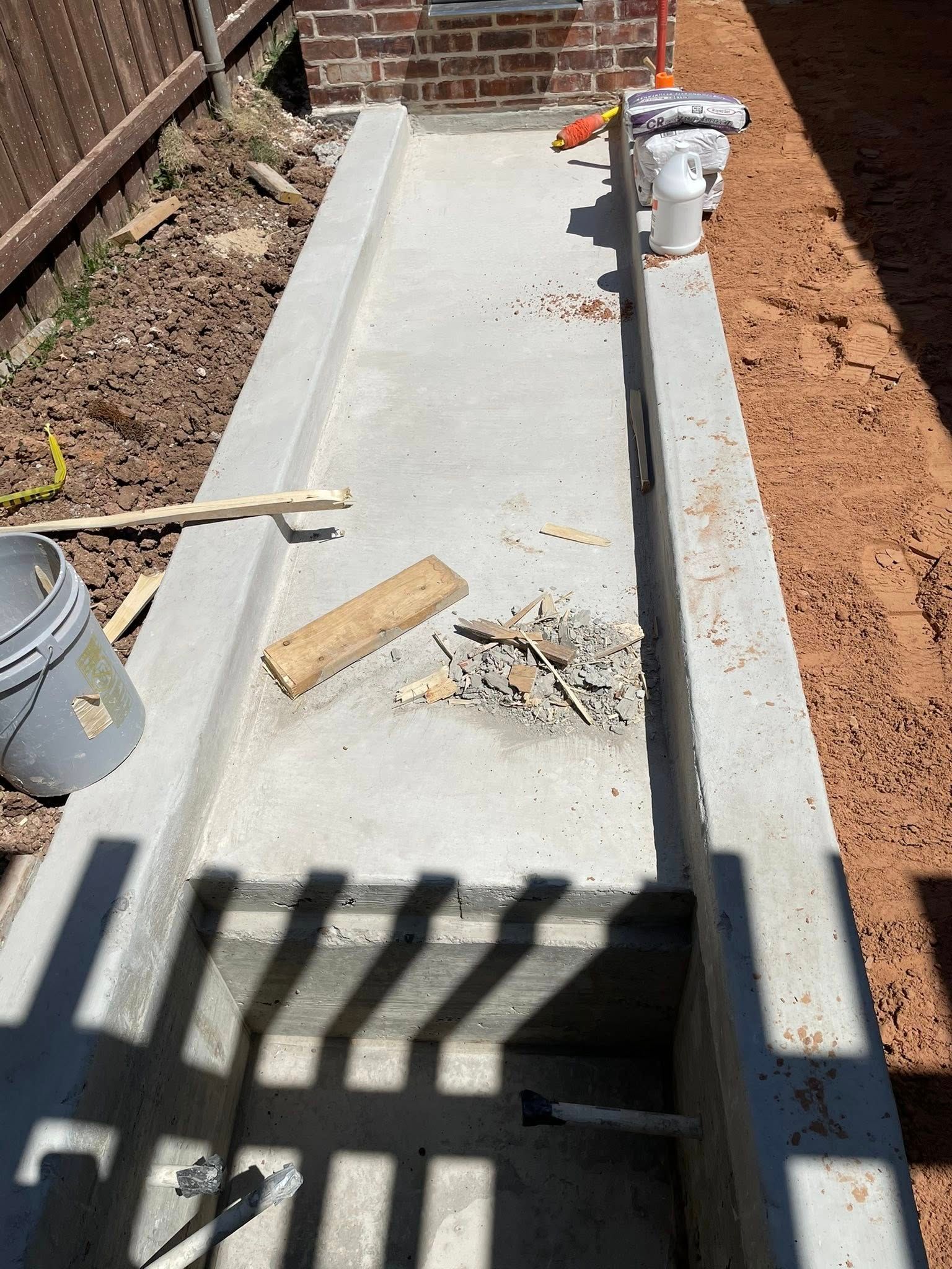 Concrete walkway under construction beside a brick wall, with tools and a pipe in a narrow trench.