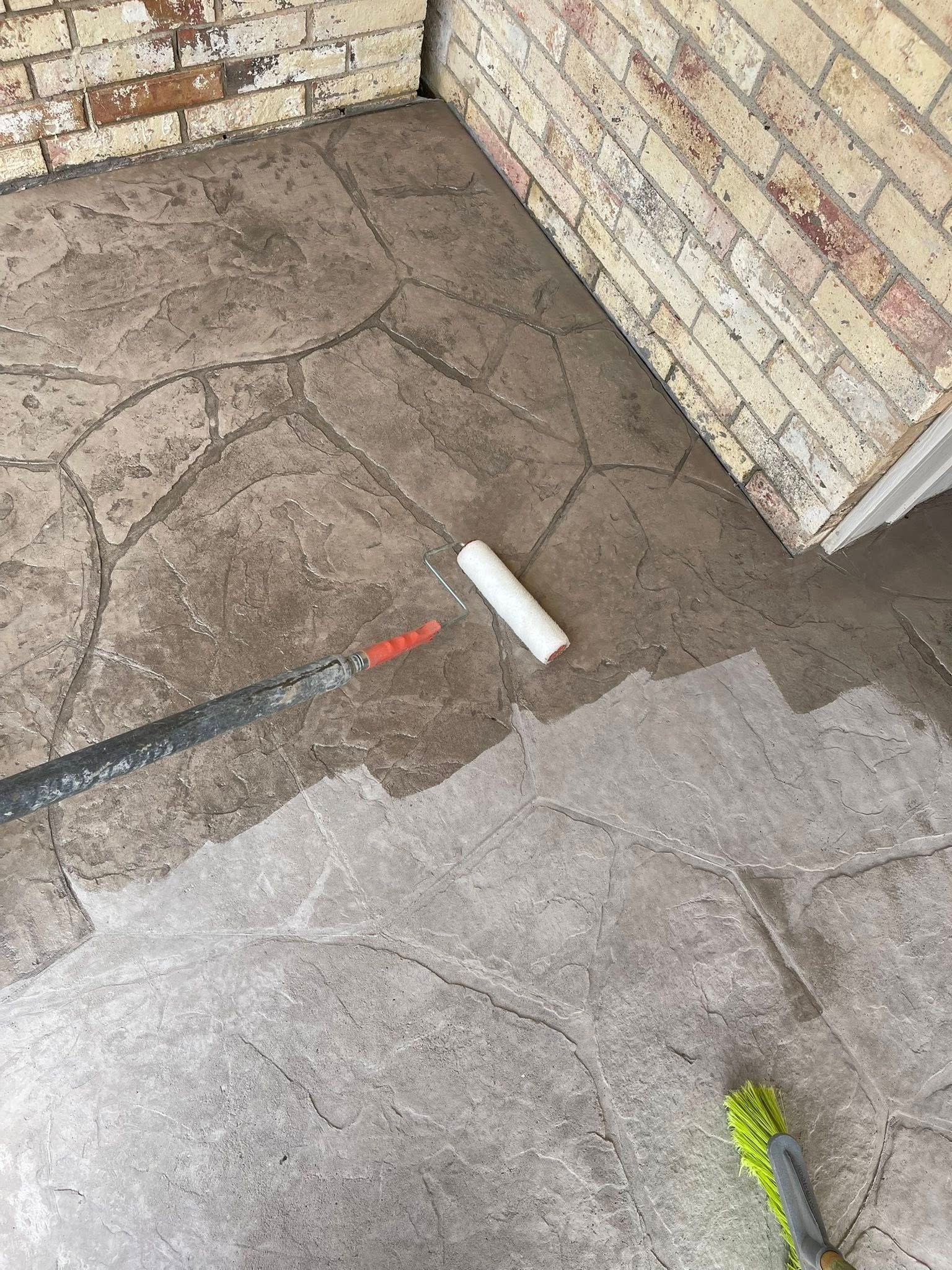 Cracked concrete porch floor beside a brick wall, with a small white pipe and a green broom at the edge.