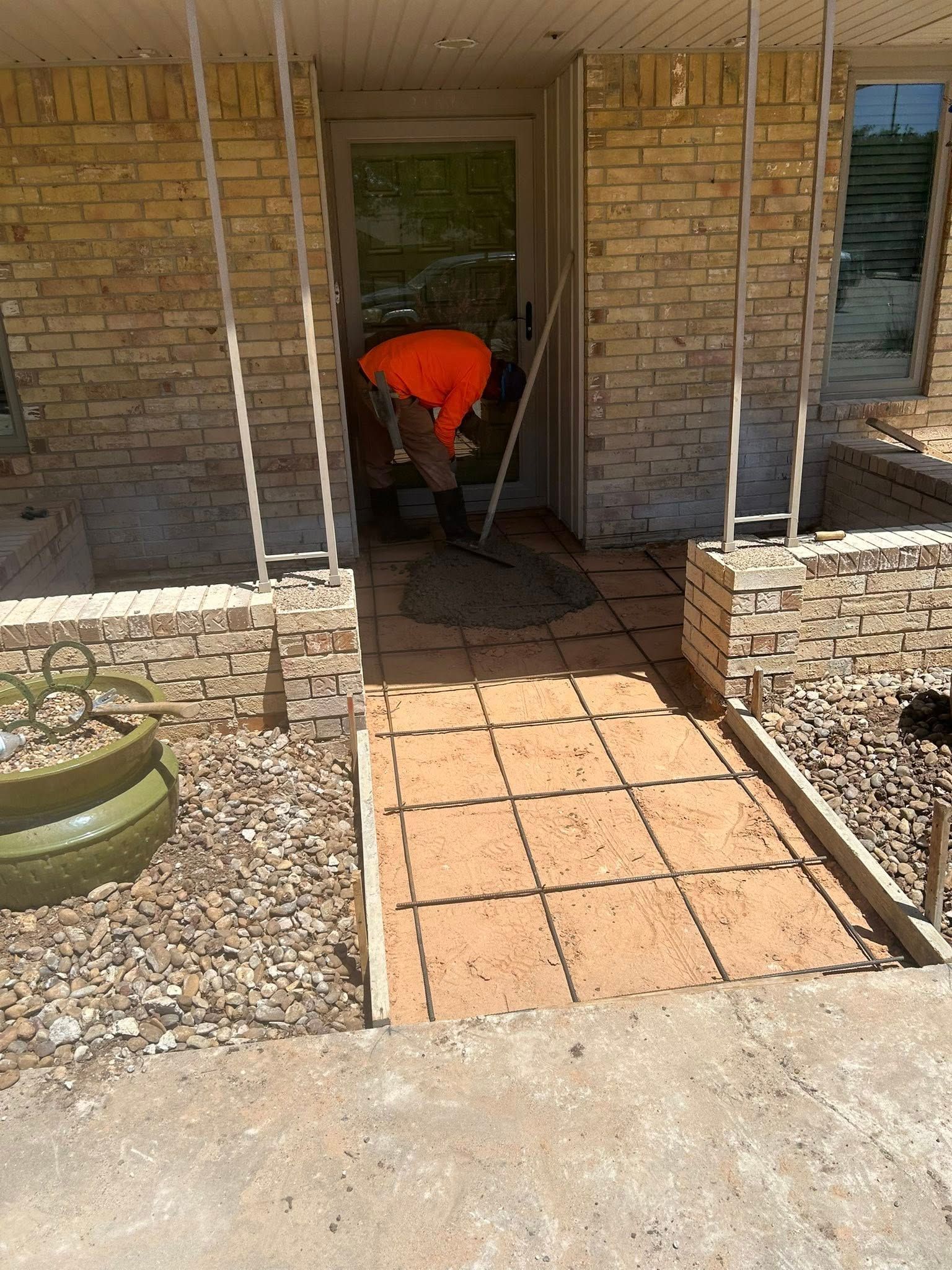Worker in orange shirt bending over a tiled walkway at a brick house entrance.