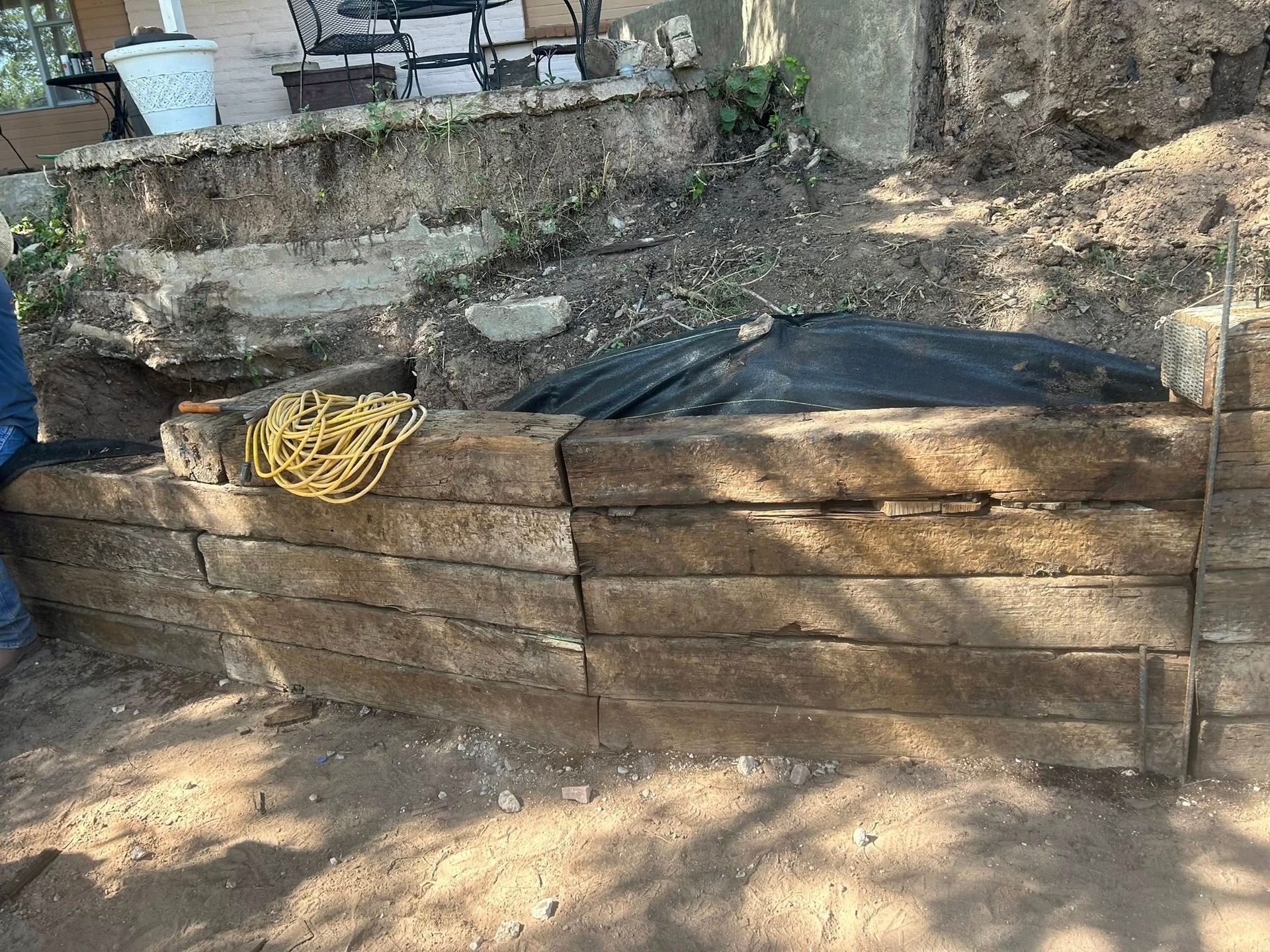 Stacked wooden retaining wall with black tarp and coiled yellow rope in a dirt yard