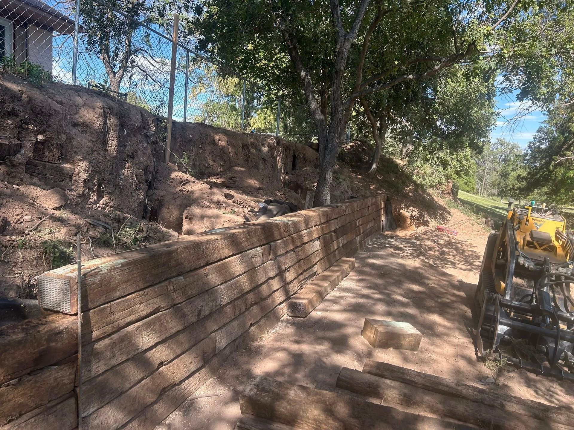 Retaining wall under construction along a wooded dirt path, with a yellow excavator on the right.