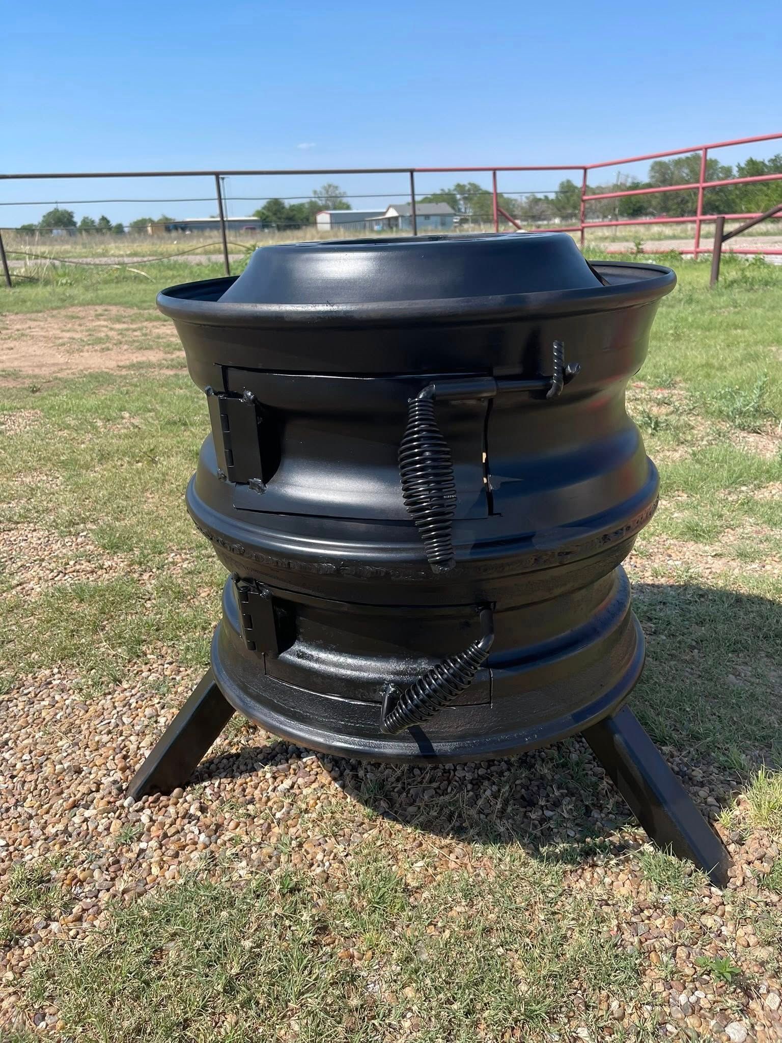 Black metal outdoor fire pit with tripod legs on gravel in a grassy field