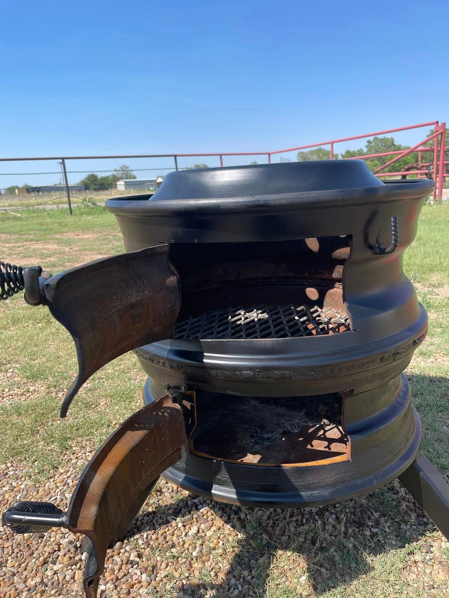 Rusty outdoor metal fire pit made from stacked drums in a gravel yard, with a blue sky behind it