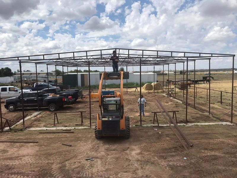 Construction site with steel frame, forklift in front, worker nearby, trucks parked on the left