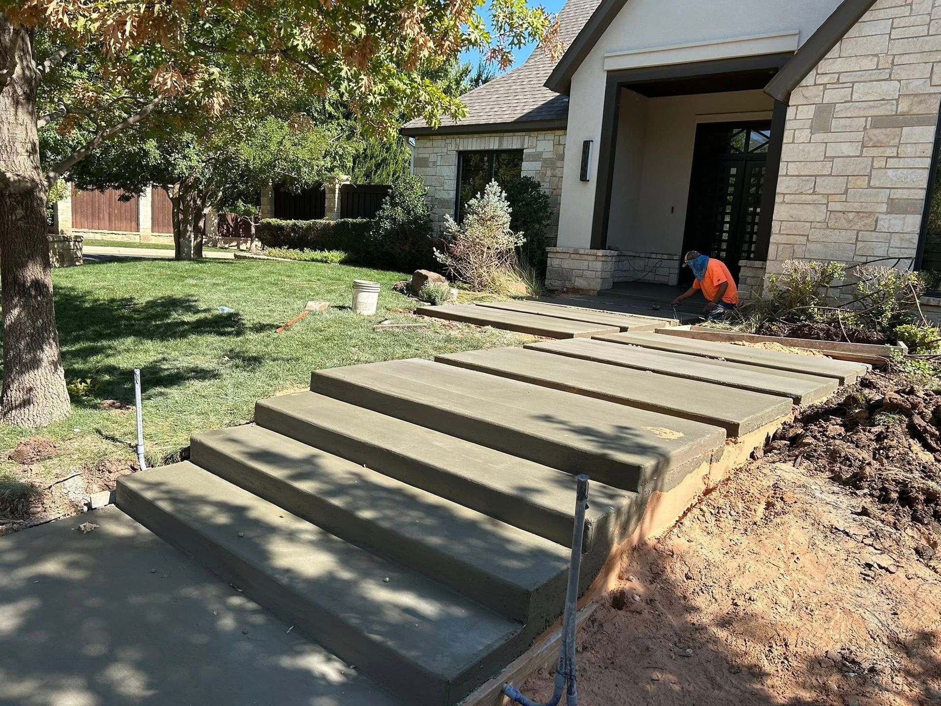 Front porch concrete steps under construction beside a stone house, with a worker kneeling nearby.