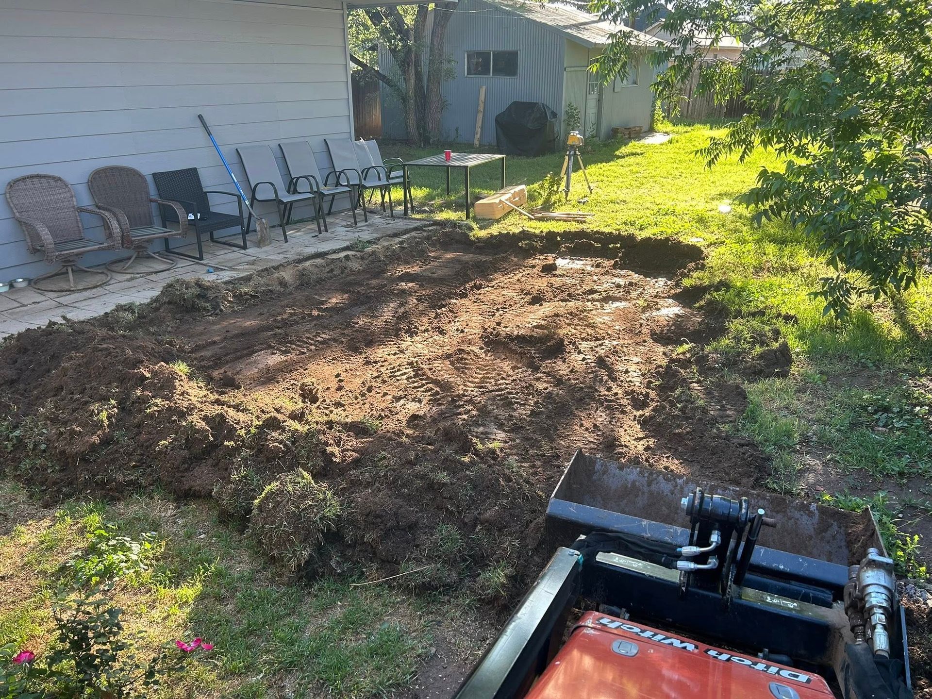 Backyard soil pile beside a house, with patio chairs lined along the wall and a lawnmower in the foreground