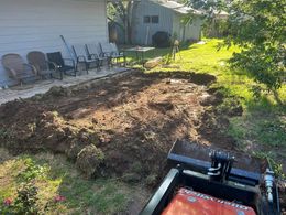 Backyard soil pile beside a house, with patio chairs lined along the wall and a lawnmower in the foreground