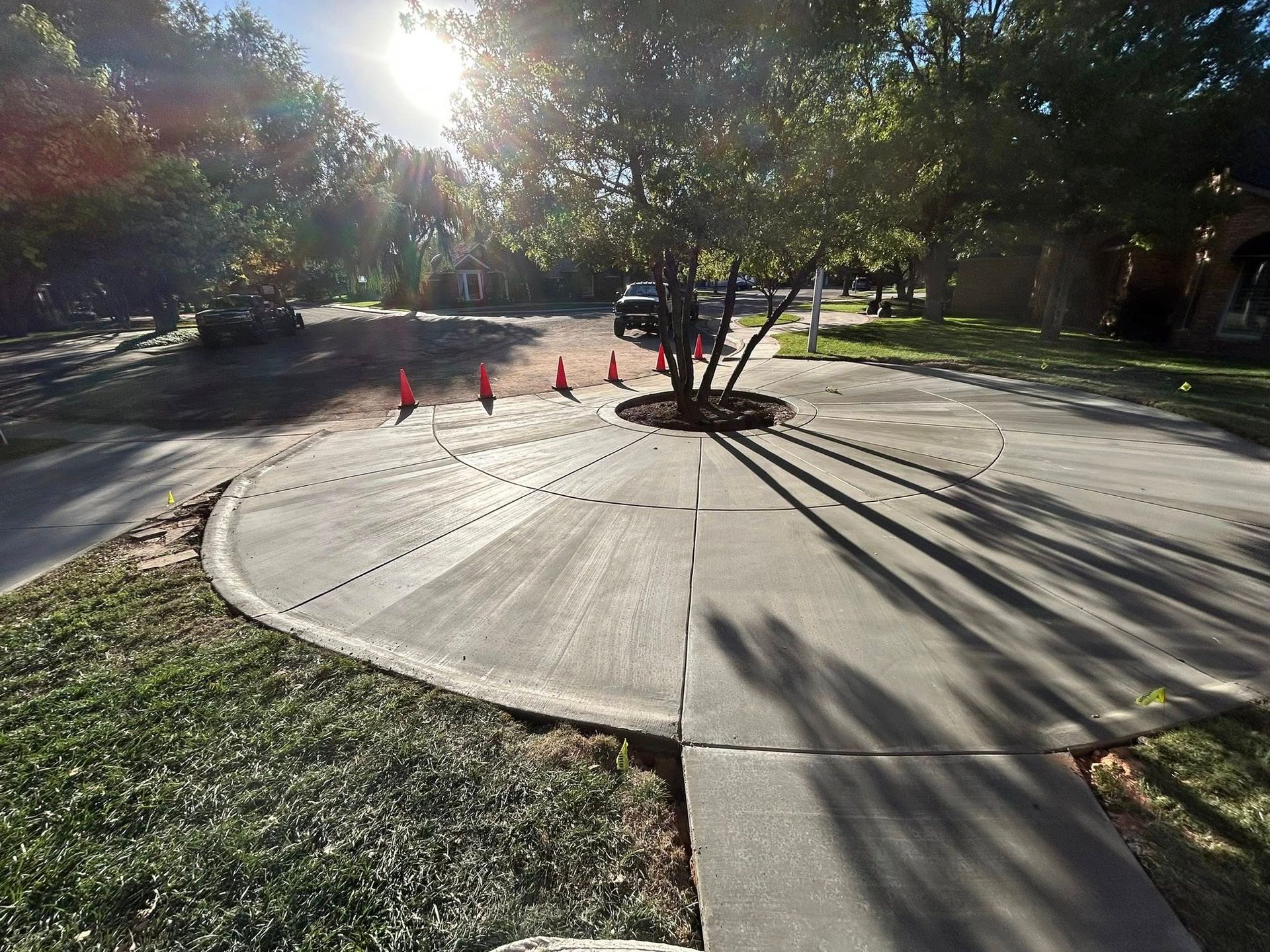 Sunlit sidewalk around a large circular concrete base with orange cones and tree shadows in a park