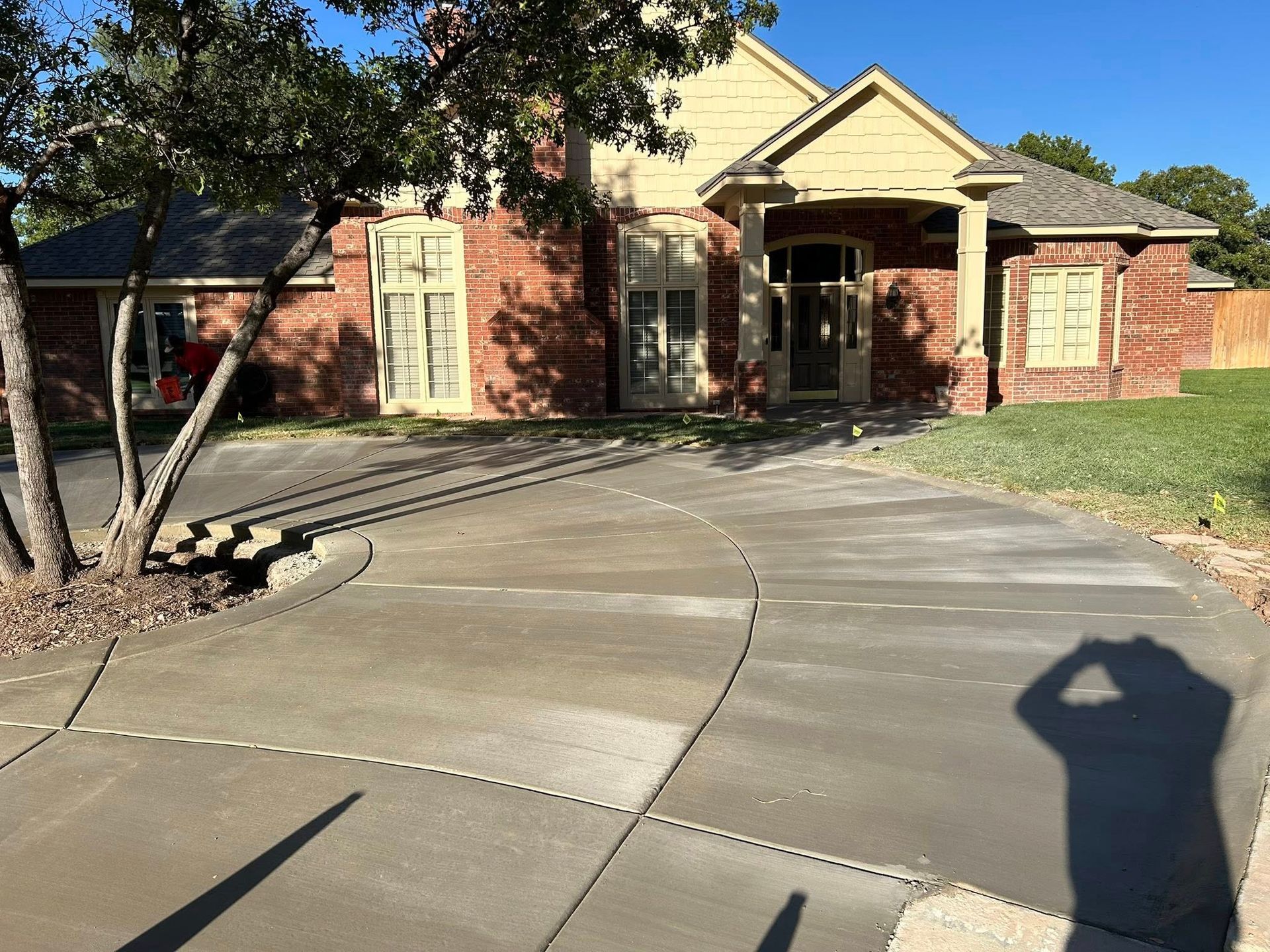 Brick house with a curved concrete driveway and a tree in the front yard, seen in bright sunlight.