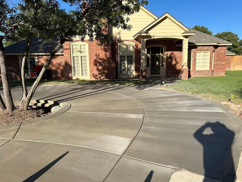 Brick house with a curved concrete driveway and a tree in the front yard, seen in bright sunlight.