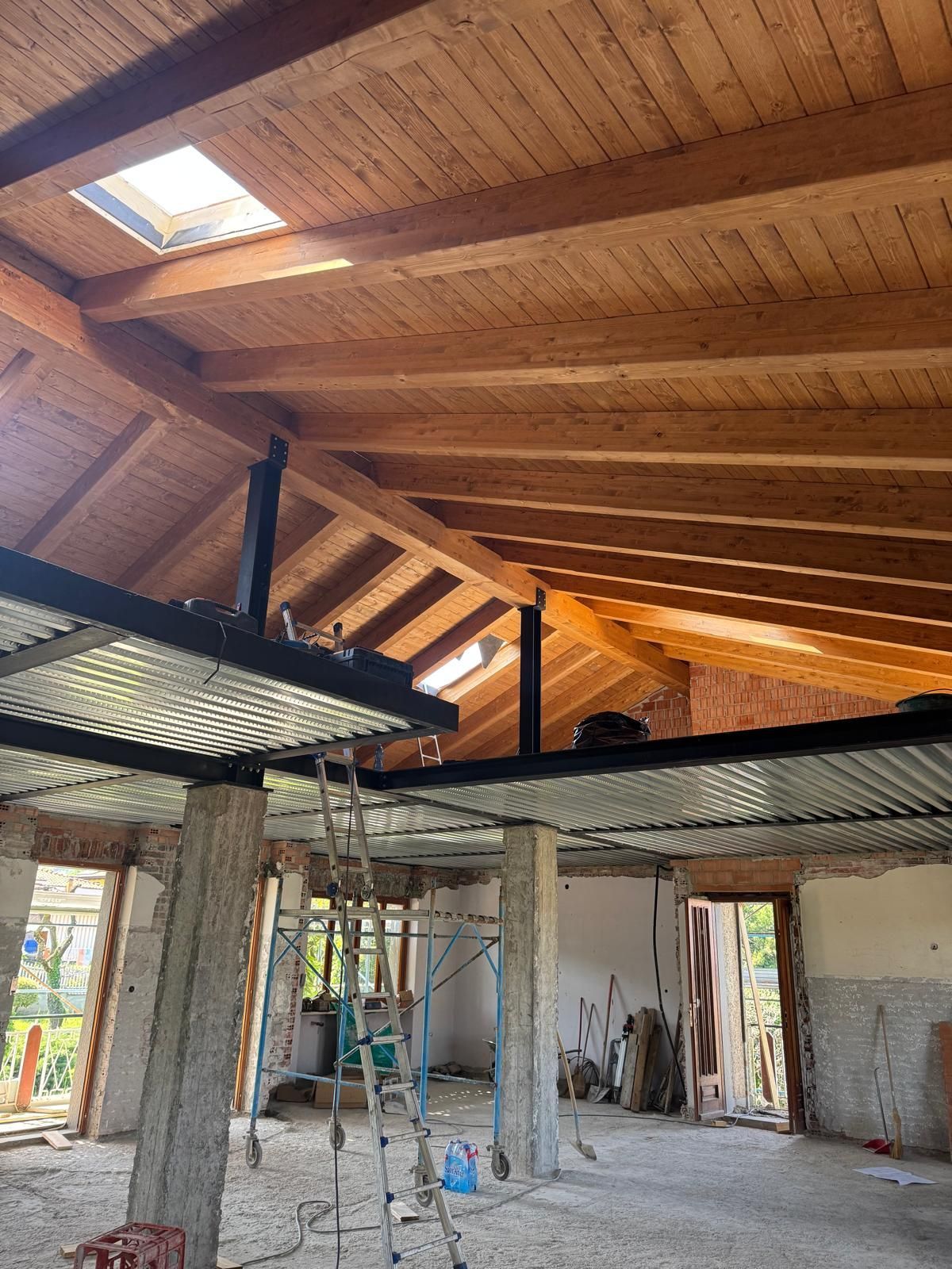 Interno in costruzione con soffitto in legno a vista, travi in metallo e pilastri in cemento.