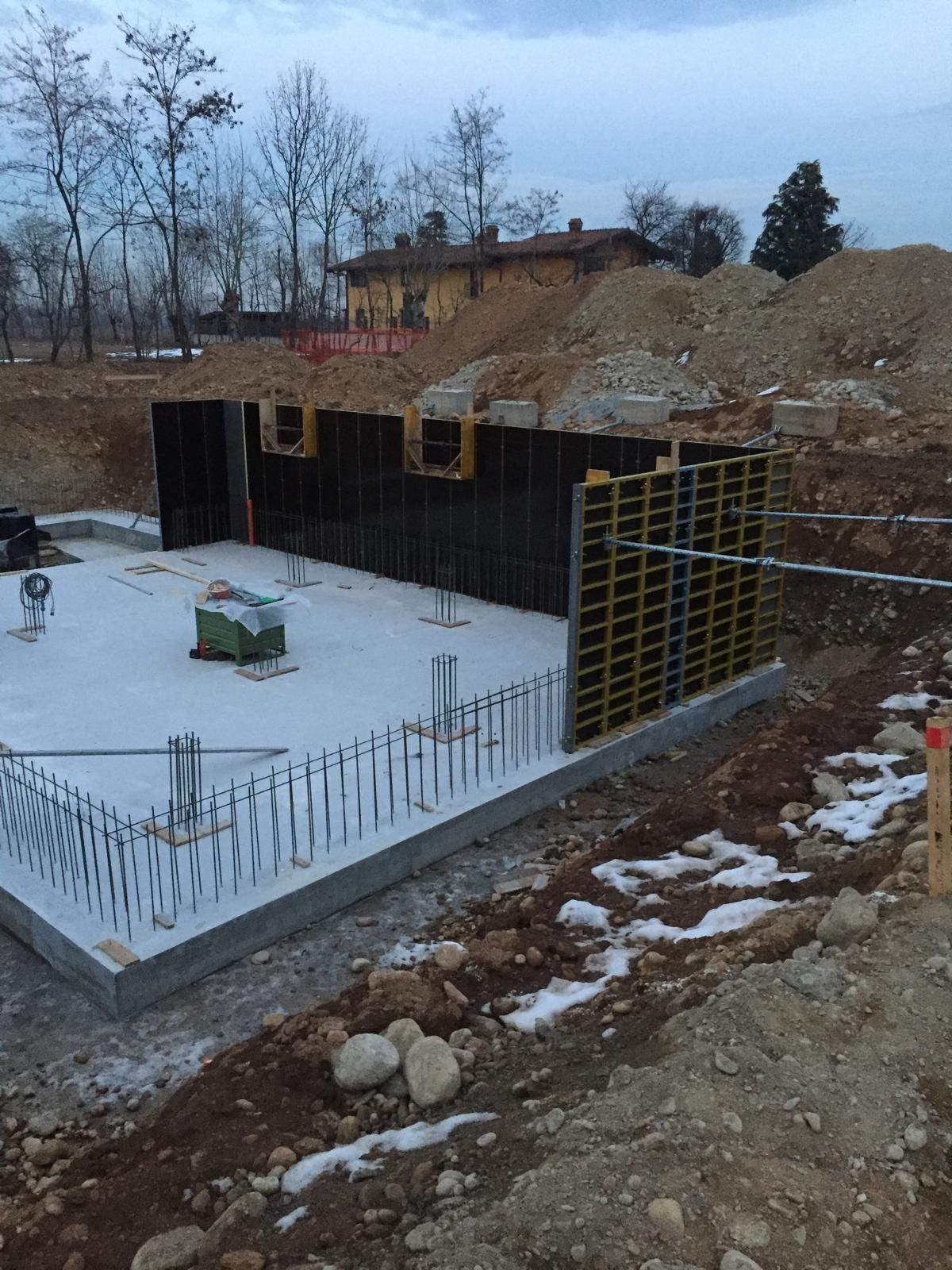 Cantiere con fondamenta, muri e casseforme in cemento. Terra e detriti circondano la struttura.