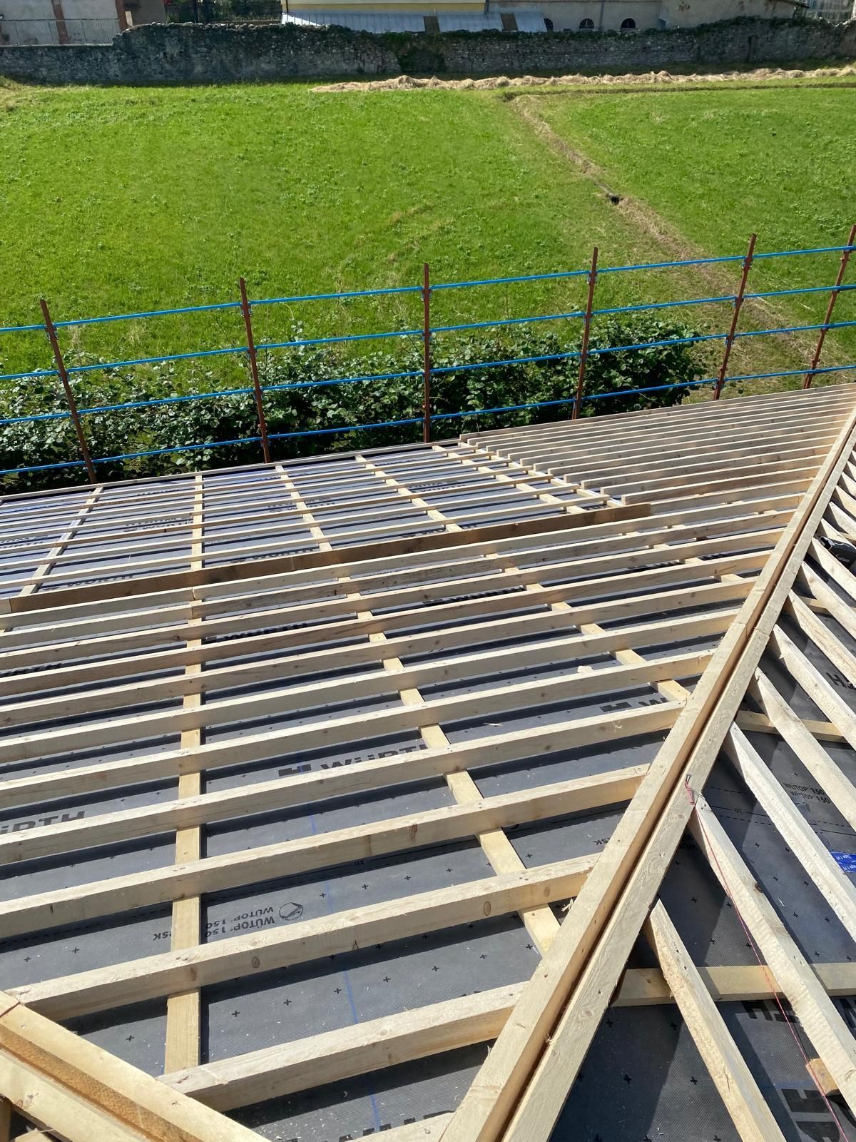 Costruzione del tetto: travi in legno, rivestimento e un sottofondo scuro; campo verde sullo sfondo.