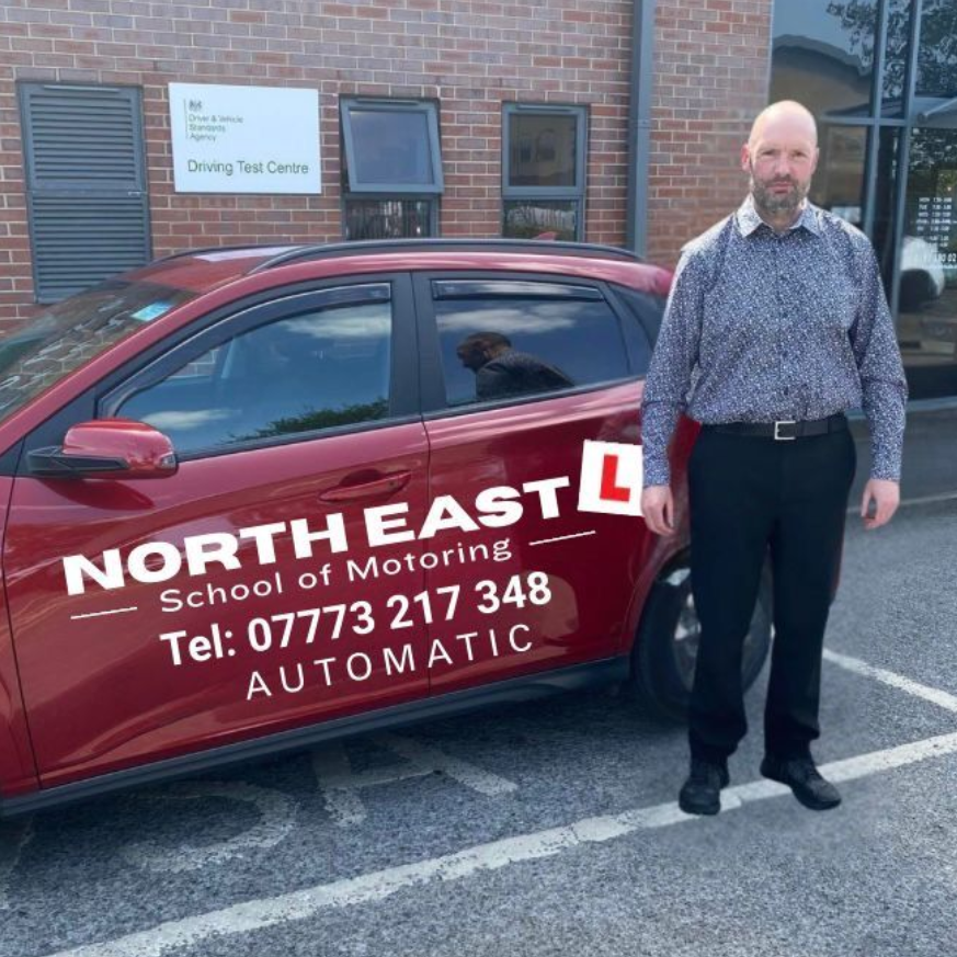 Man standing beside a maroon North East School of Motoring car outside a brick building.