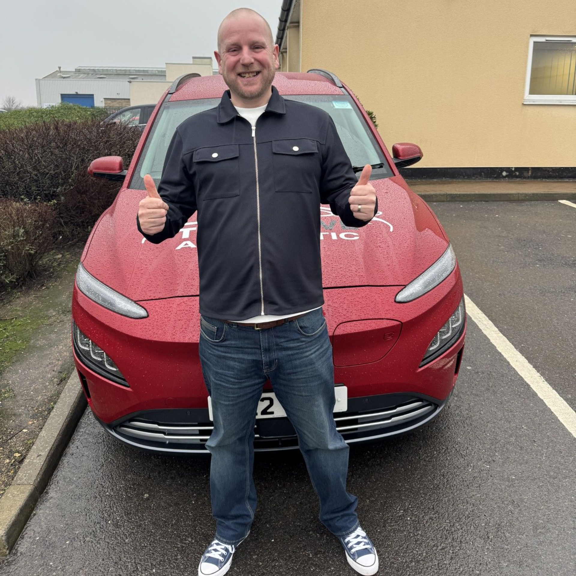 Man standing in front of a red car, smiling and giving two thumbs up in a parking lot.