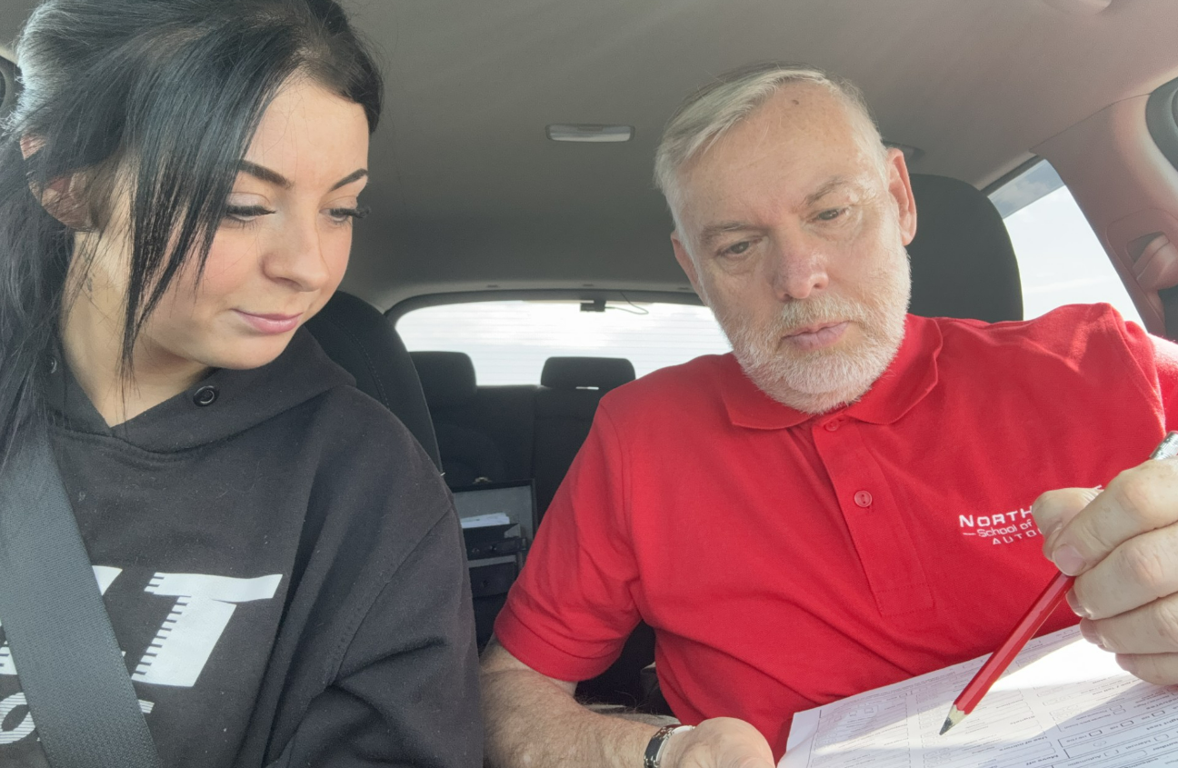Two people in a car reviewing paperwork, one holding a pen and looking down at a form.