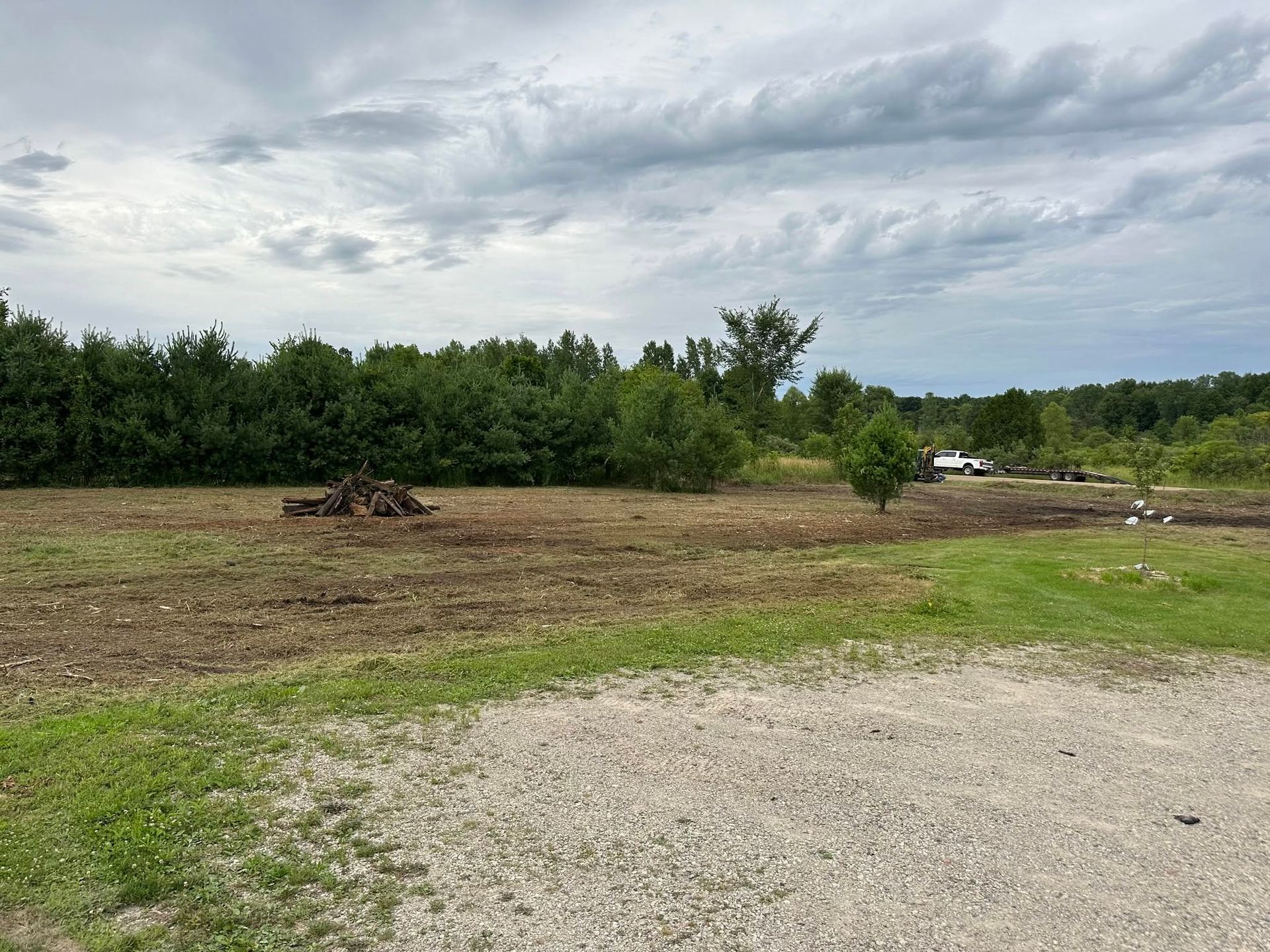 Gravel foreground with grassy field, dense green trees, and a cloudy sky over open land