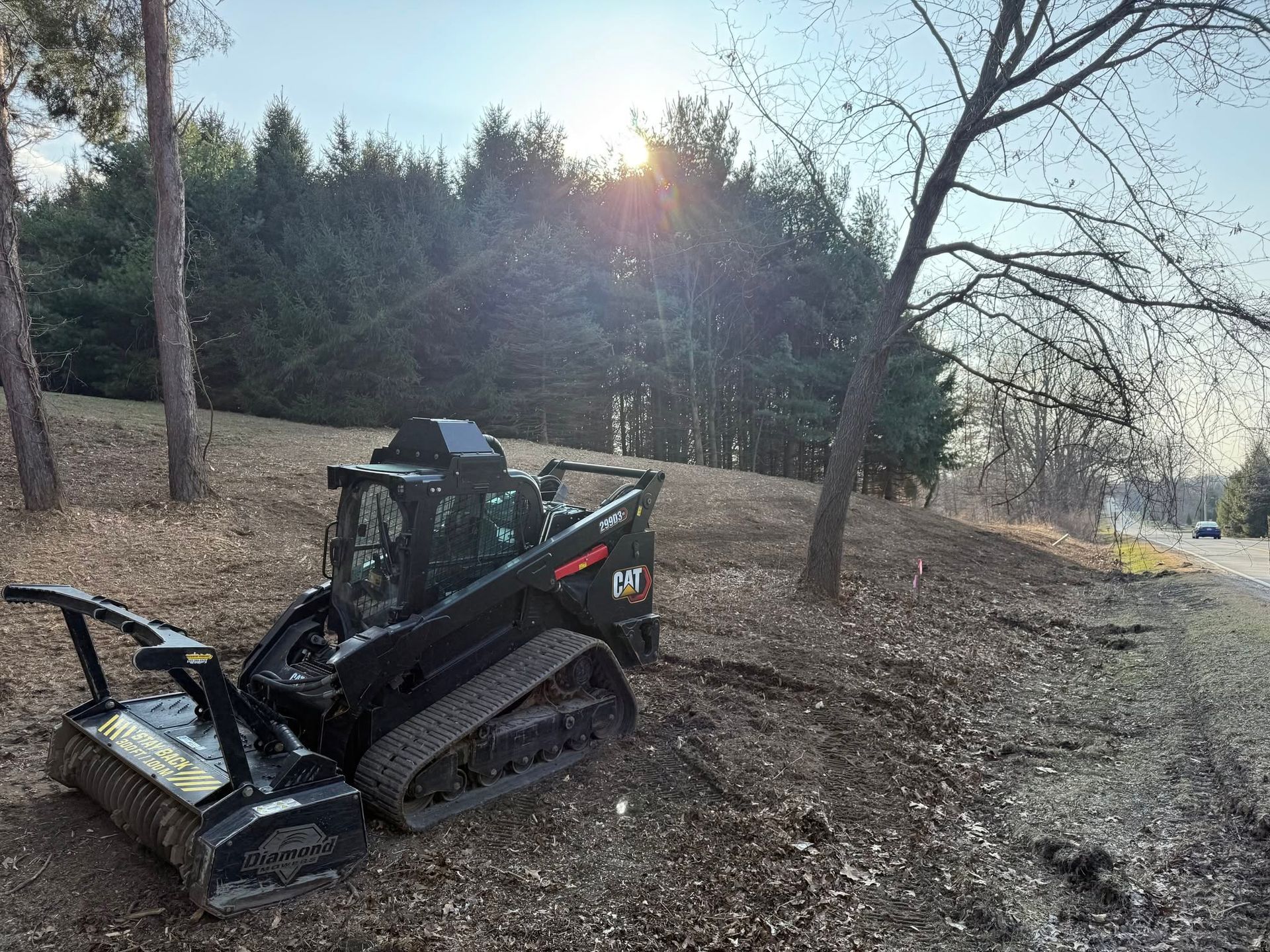 Black skid steer with brush cutter on a wooded hillside at sunset