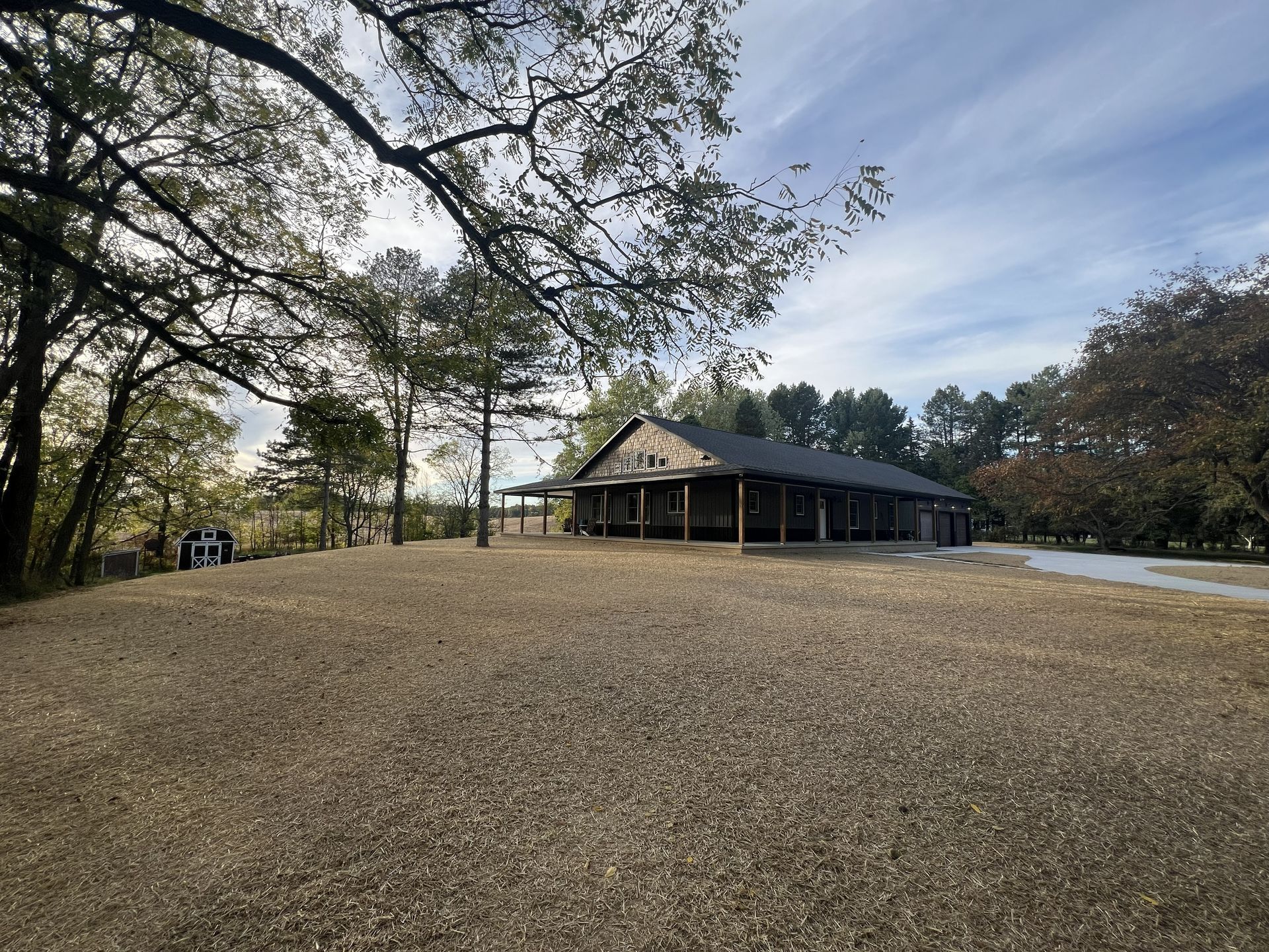 Wide gravel yard with a rustic pavilion among trees under a cloudy sky.