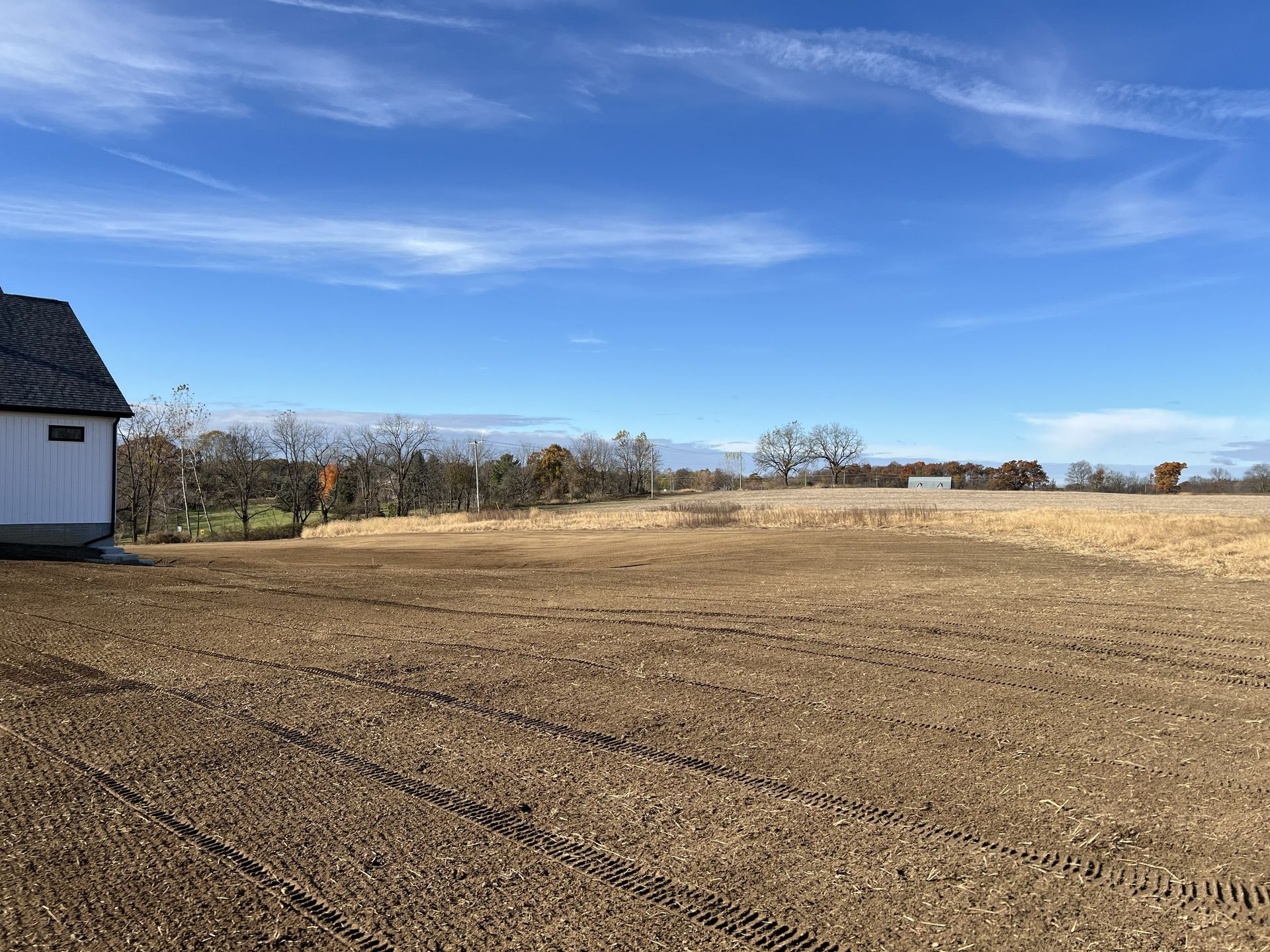 Plowed field under a blue sky beside a white house and bare trees