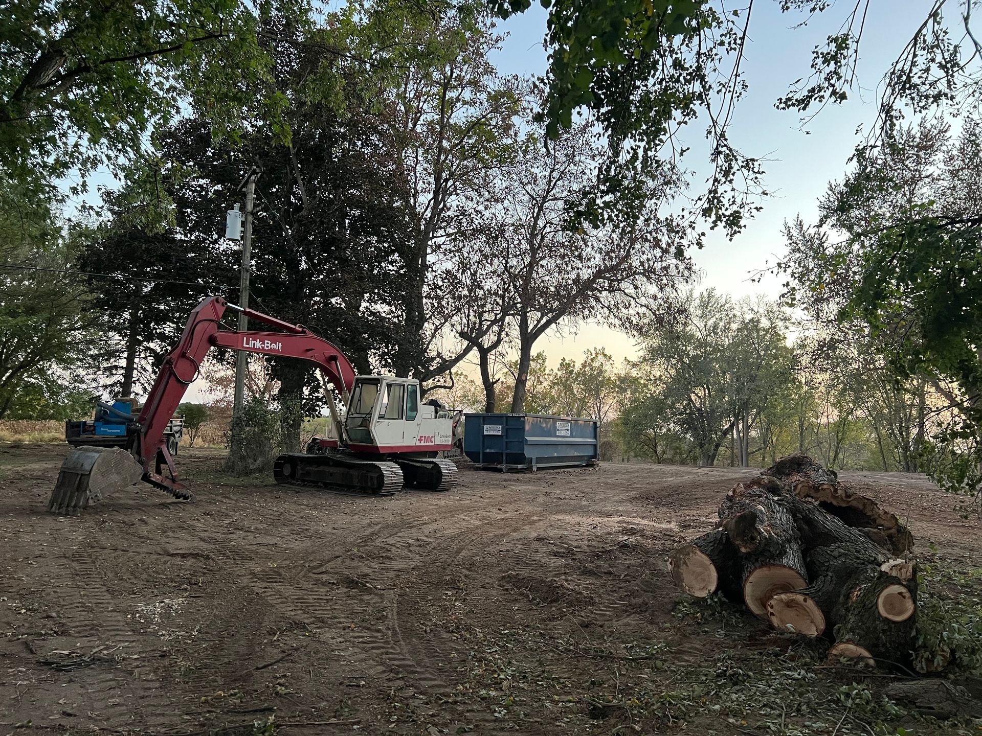 Excavator working in a wooded dirt clearing beside a blue trailer, with cut logs stacked nearby.