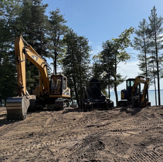 Two excavators and a compact loader on a dirt site among pine trees under a blue sky