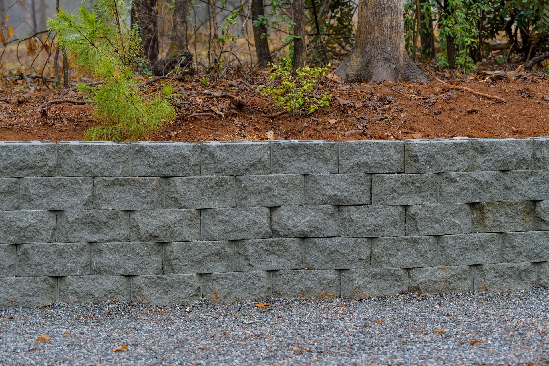 Gray stone retaining wall beneath a wooded slope with gravel foreground in autumn colors