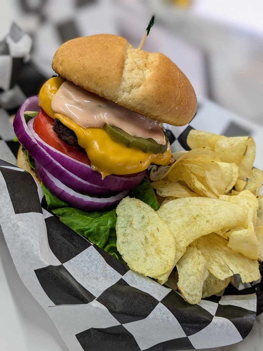 A hamburger with cheese , onions , tomatoes and potato chips on a checkered paper.