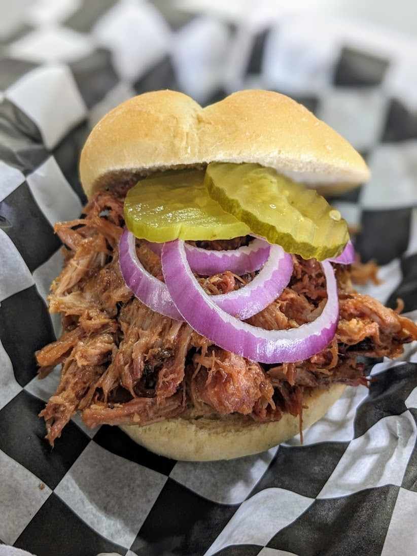 A pulled pork sandwich with onions and pickles on a bun on a checkered paper.