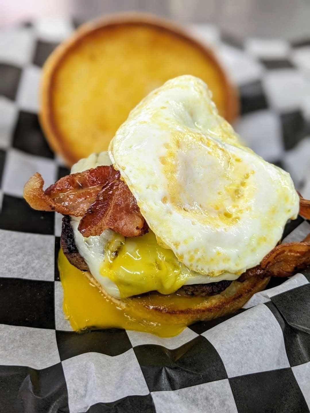 A close up of a hamburger with eggs , bacon and cheese on a checkered paper.