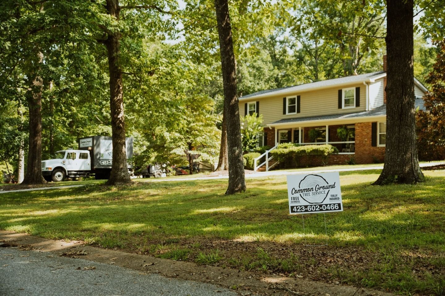 White two-story house among tall trees with a yard sign and a truck parked nearby