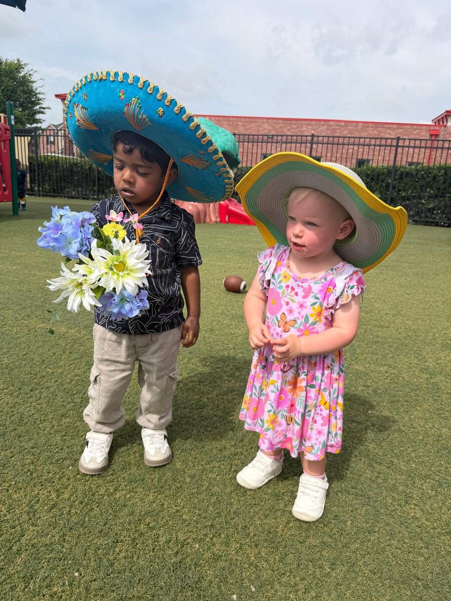 Two children in sombreros stand on grass, one holding flowers, at an outdoor event.