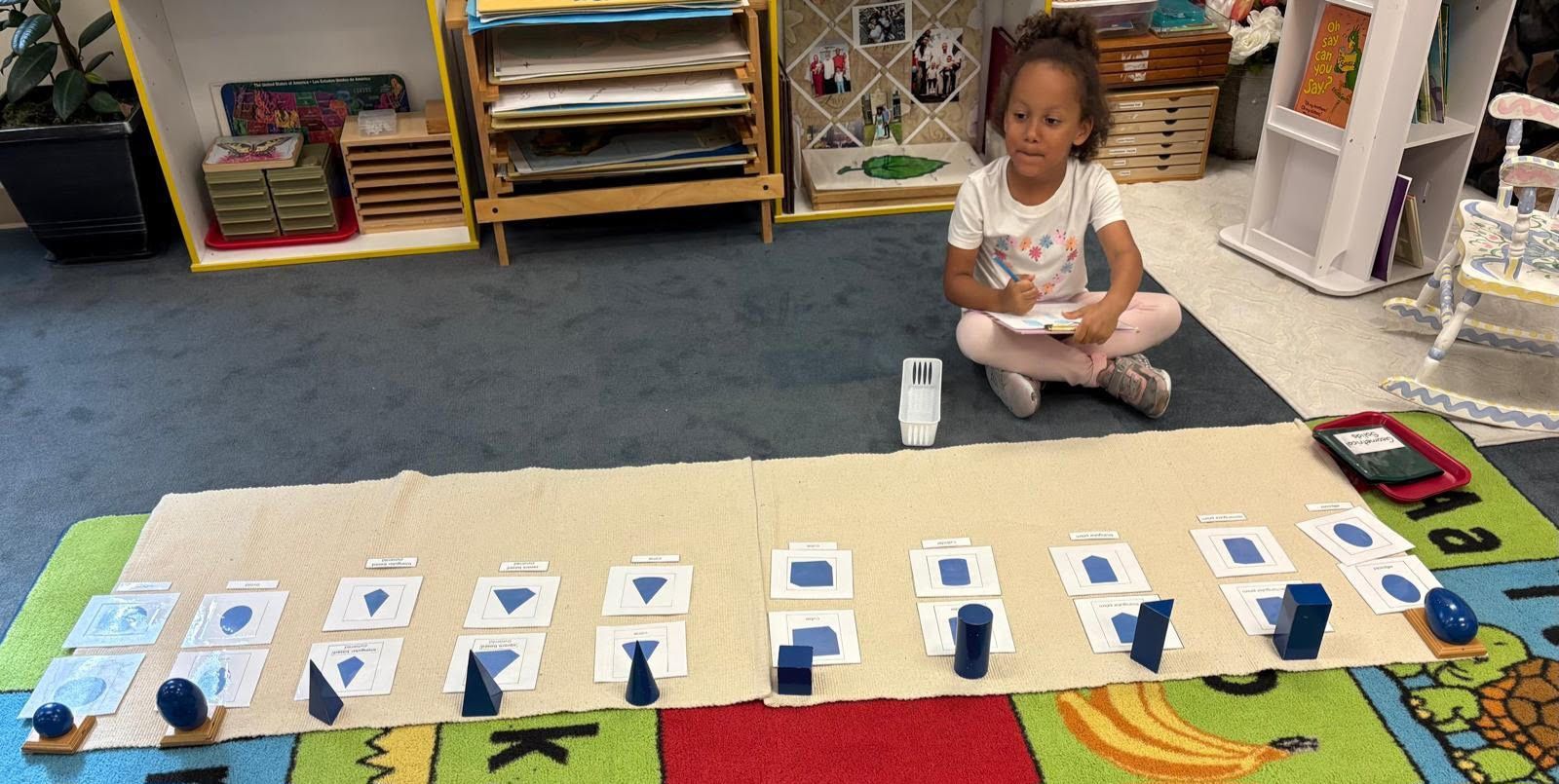 A primary Montessori student identifies and matches blue geometric solids to their corresponding 2D bases and nomenclature cards on a long floor mat.