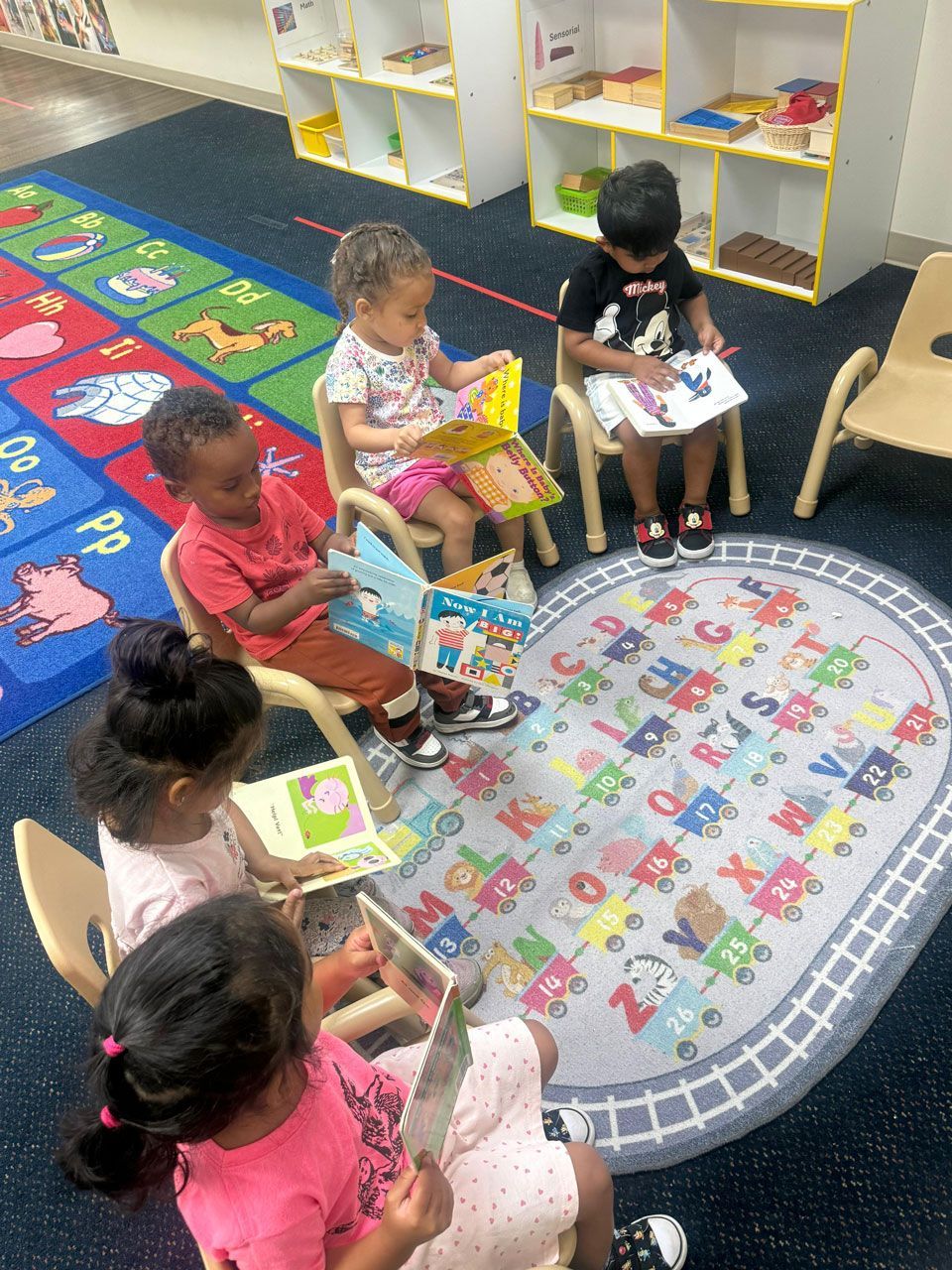 Toddler Montessori children sit in a circle on small chairs, independently reading board books on an alphabet rug.