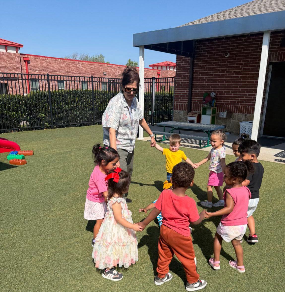 A Montessori teacher and a group of toddlers hold hands in a circle for an outdoor song and movement activity.
