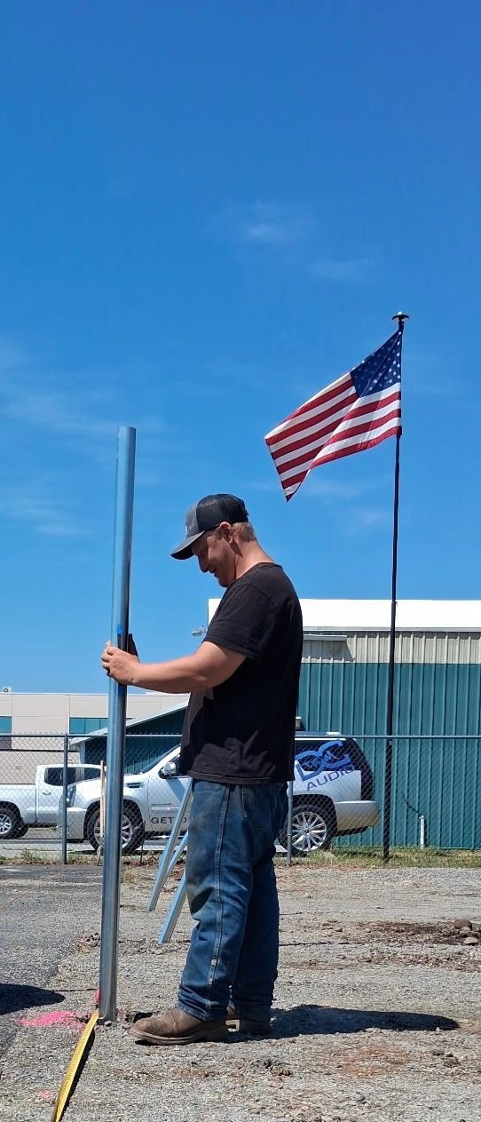 Man standing beside a fence holding a pole, with an American flag flying against a blue sky.
