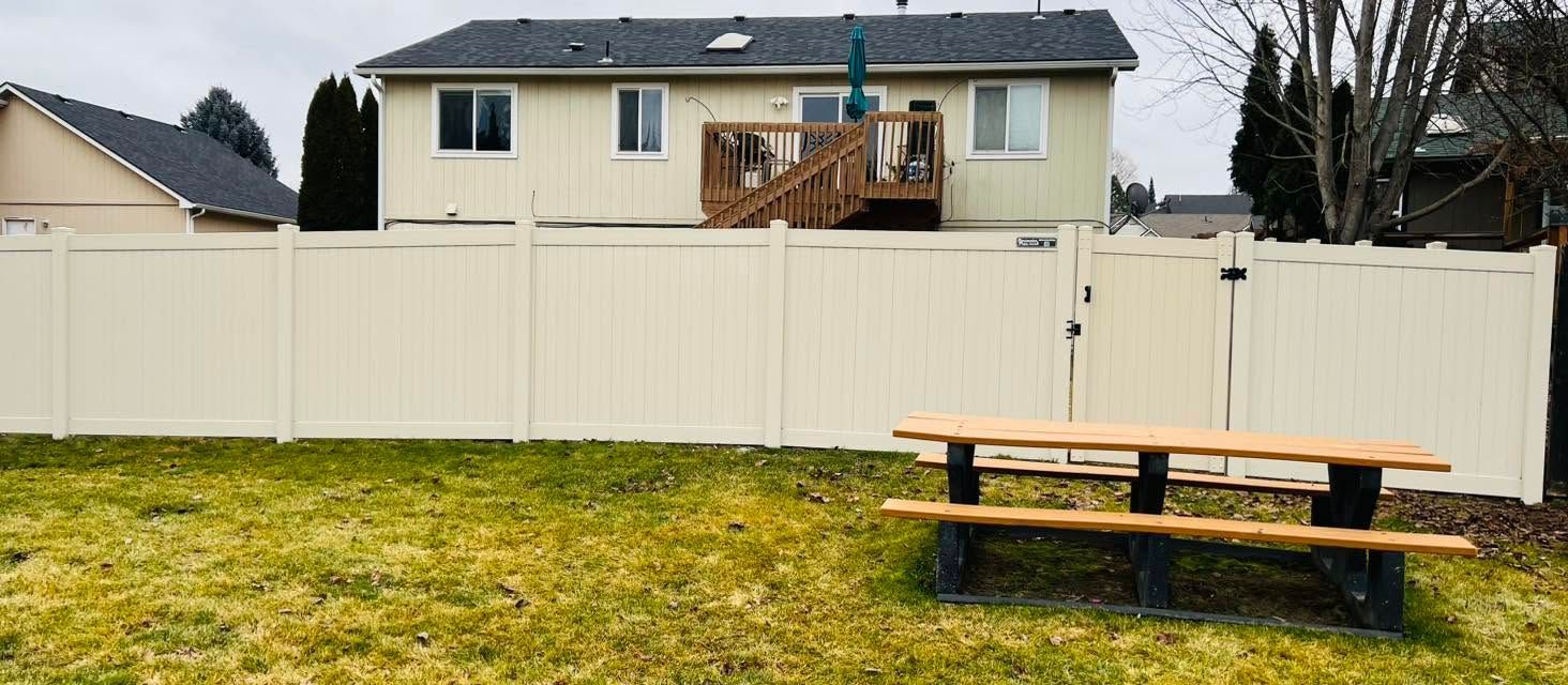 Backyard with a beige house, white fence, and a wooden picnic table on grass.