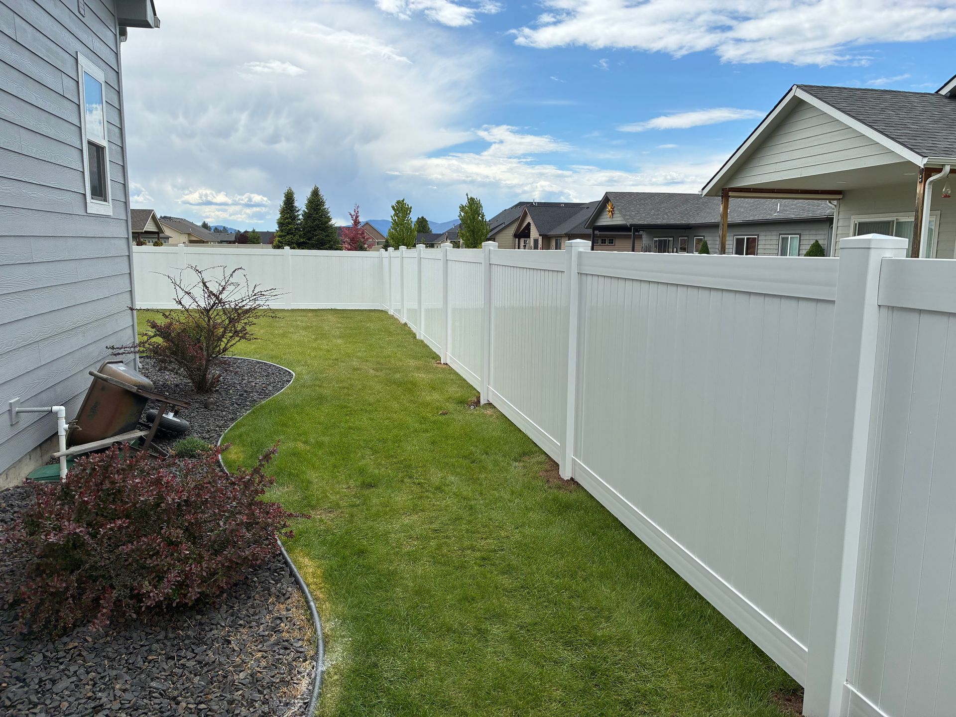 Side yard with green grass, white vinyl fence, and gray house exterior under a cloudy sky