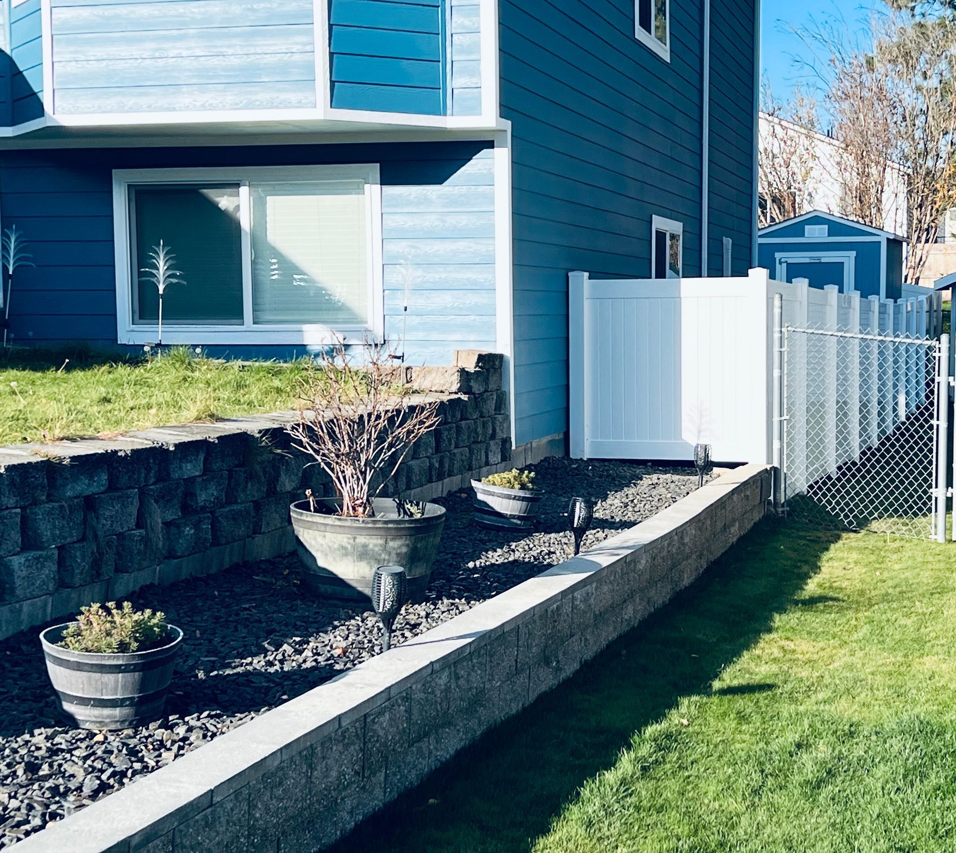 Blue townhouse exterior with raised stone garden bed, potted plants, and a white fence along a grassy yard