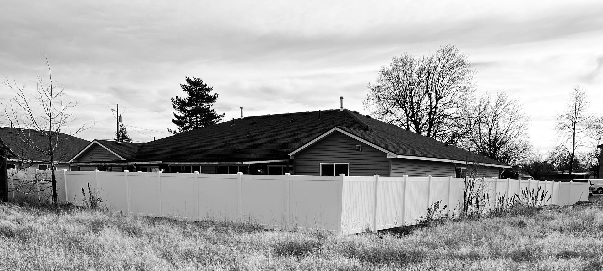 Black-and-white view of a suburban house behind a white fence under a cloudy sky