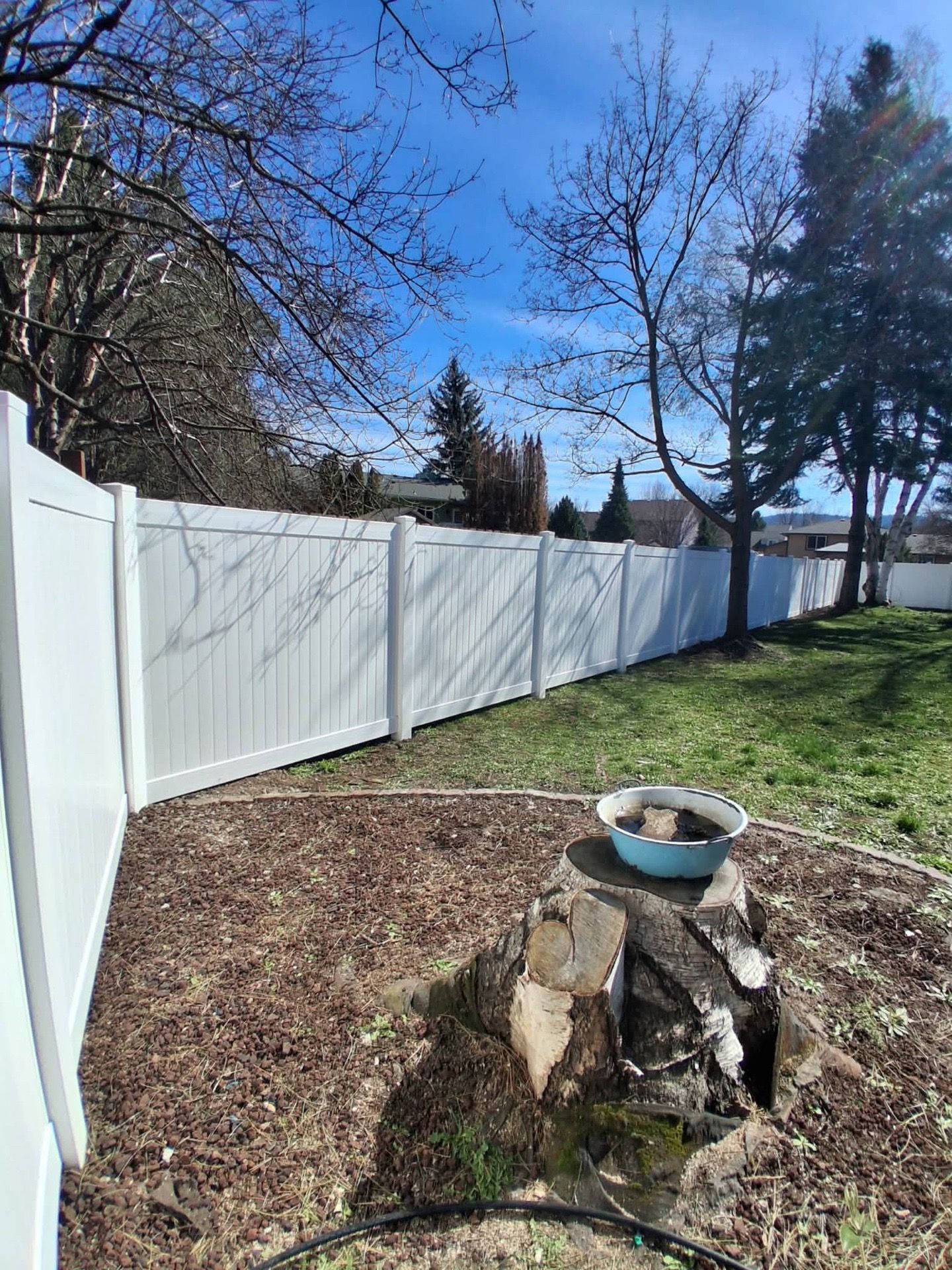 Backyard with white fence, bare trees, and a stone fire pit on mulch in sunny weather