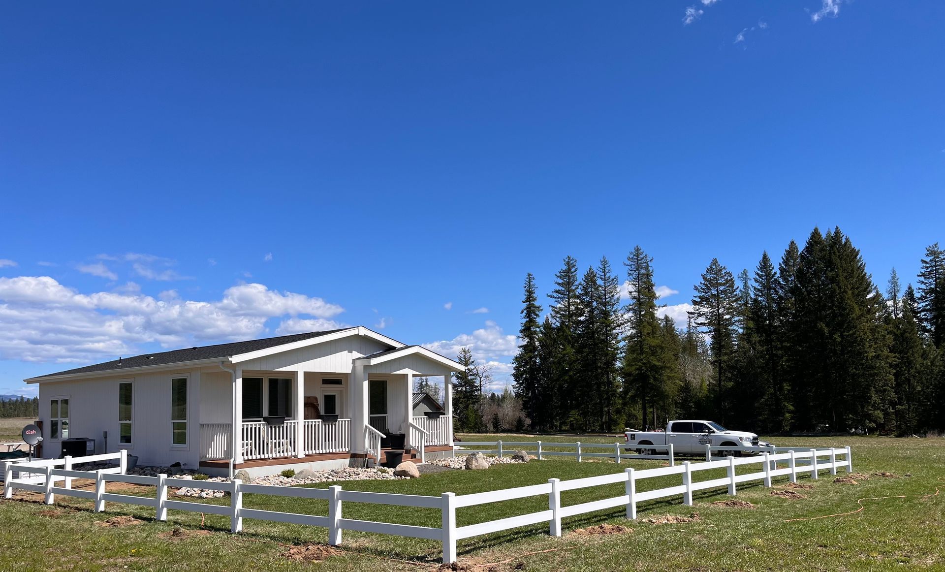 White farmhouse with a porch and white fence in a grassy field under a blue sky, with trees and a parked truck.