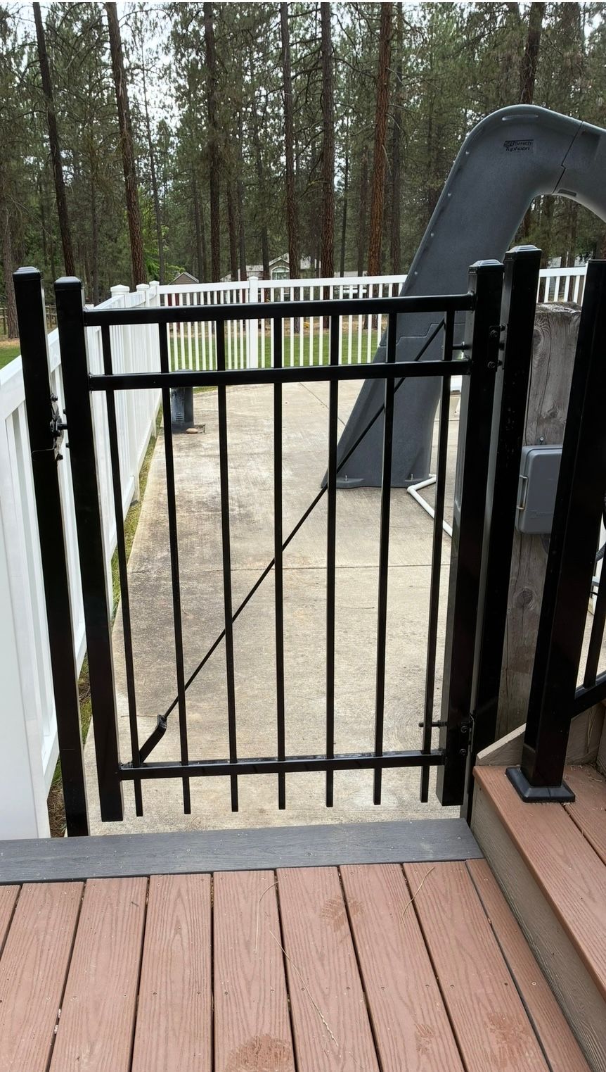 Black metal gate on a wooden deck overlooking a gravel yard and trees beyond.