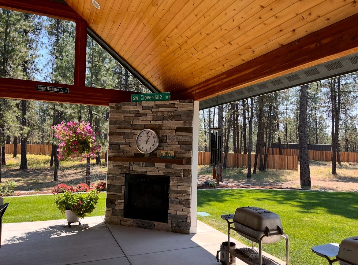 Covered patio with stone fireplace, wood ceiling, and grassy yard with trees beyond